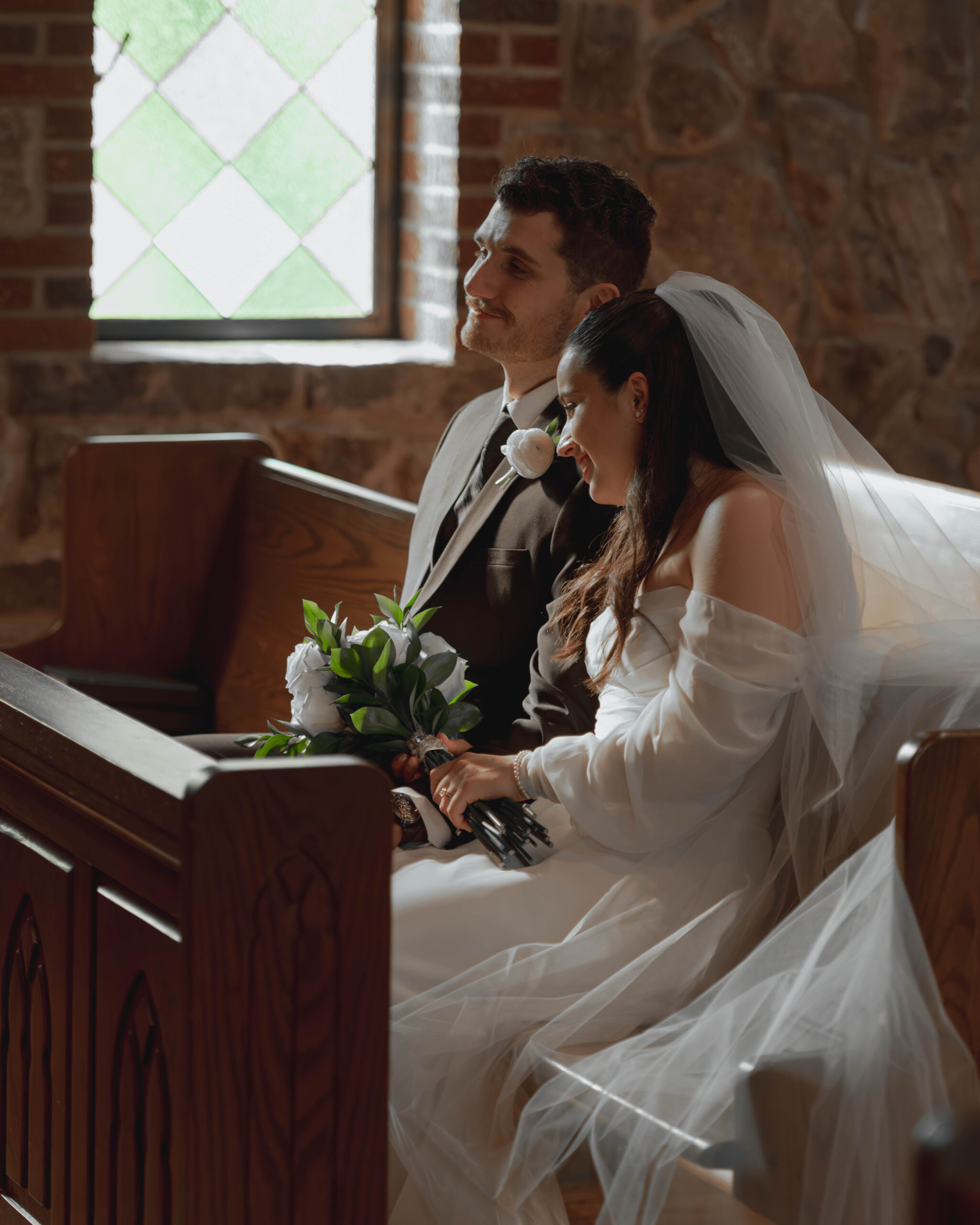 A couple who is just married sits on a pew in front of a stained glass window smiling.