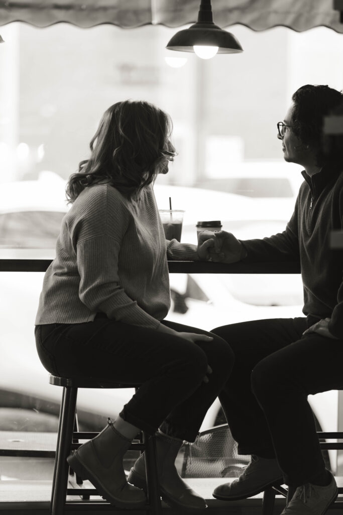 A man and woman sit on barstools at a coffee shop during their engagement session and hold hands while staring out the window.