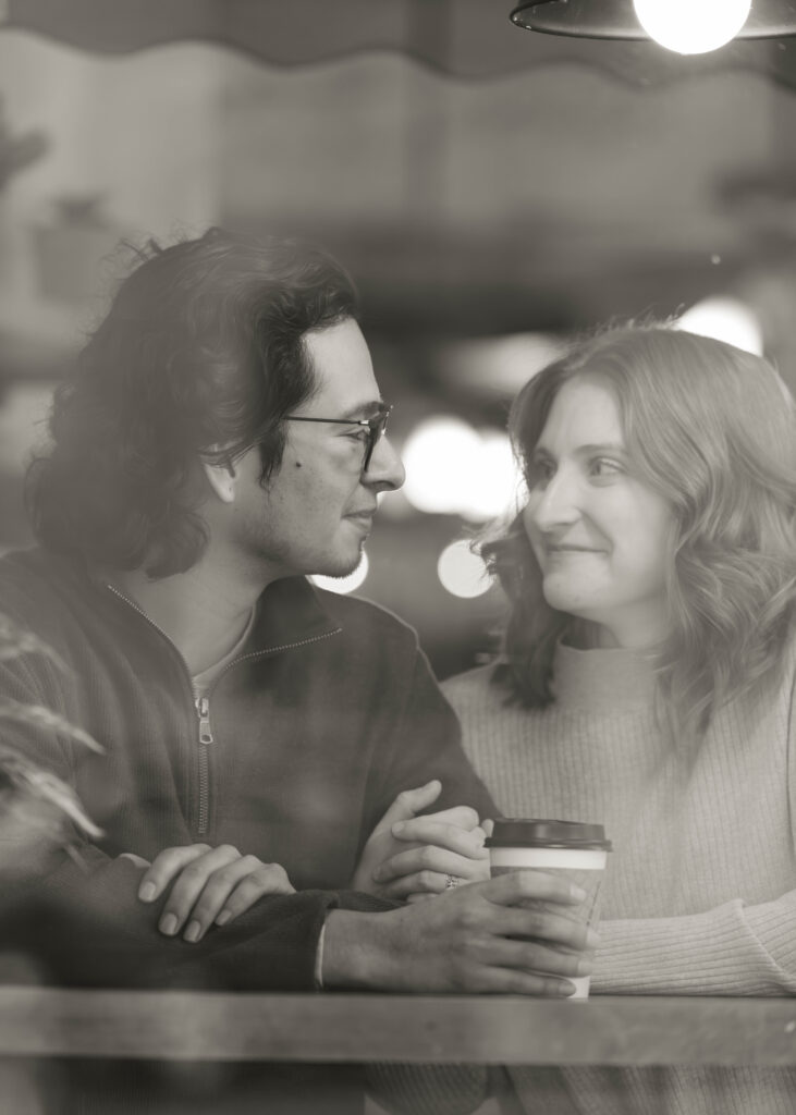 A man and woman sit behind a reflective window during their engagement session and smile at each other.