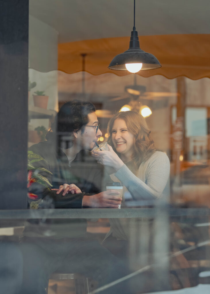 A man and woman sit behind a reflective window during their engagement session. The man is sipping the woman's coffee wit a funny face and she is laughing.