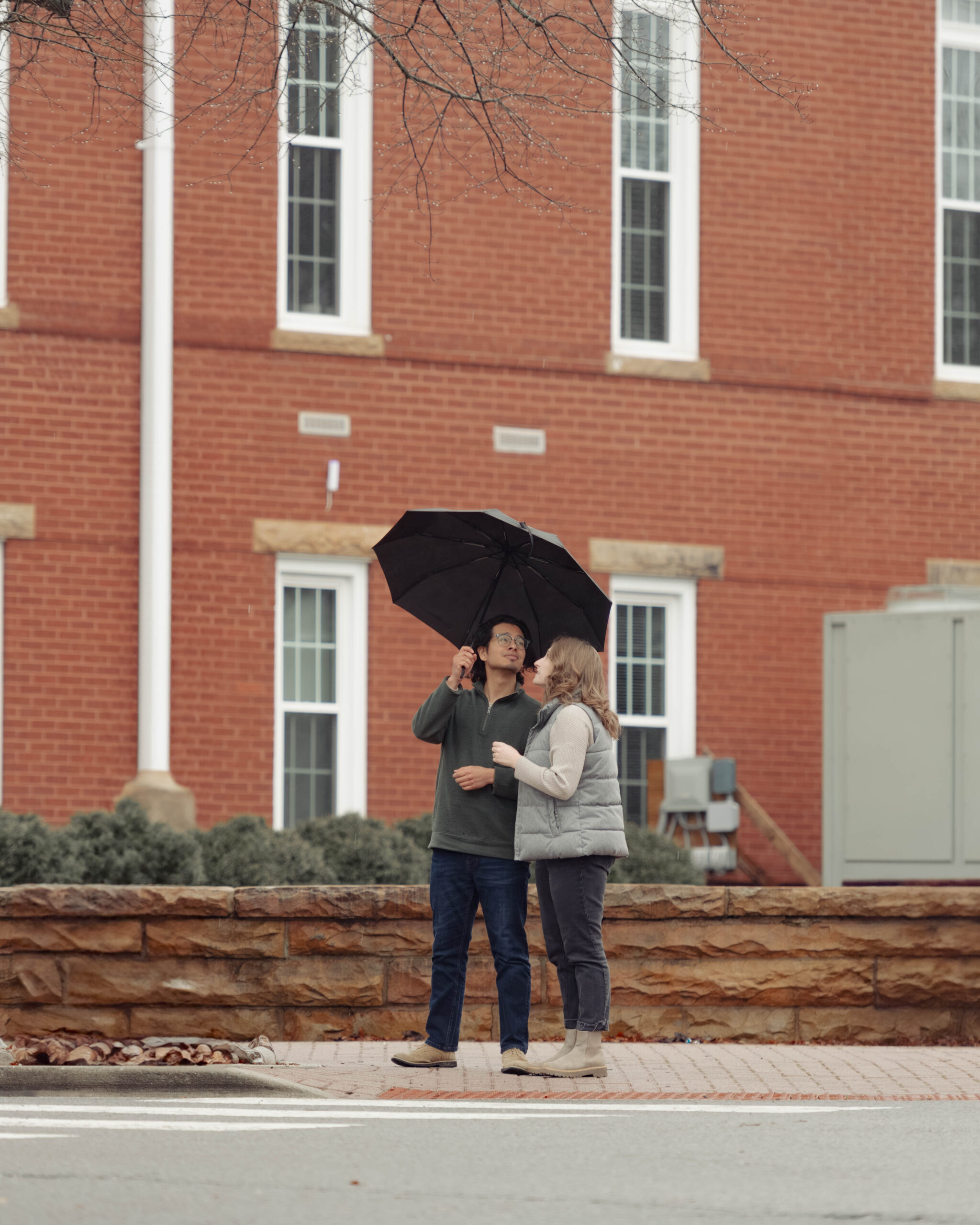 A man and woman stand on a street corner under an umbrella during their engagement session.