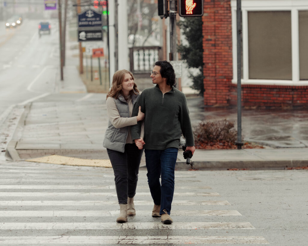 A man and woman cross a rainy street during their engagement session. The woman is hugging onto the man's arm and they are smiling at each other