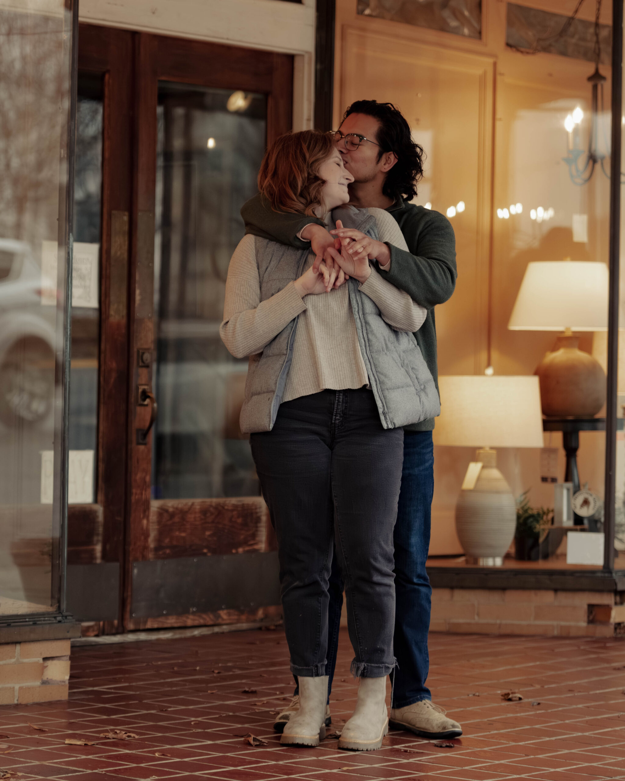 A man hugs a woman from behind and kisses her forehead during their engagement session. They are standing in front of a lamp store. Their fingers are laced together.