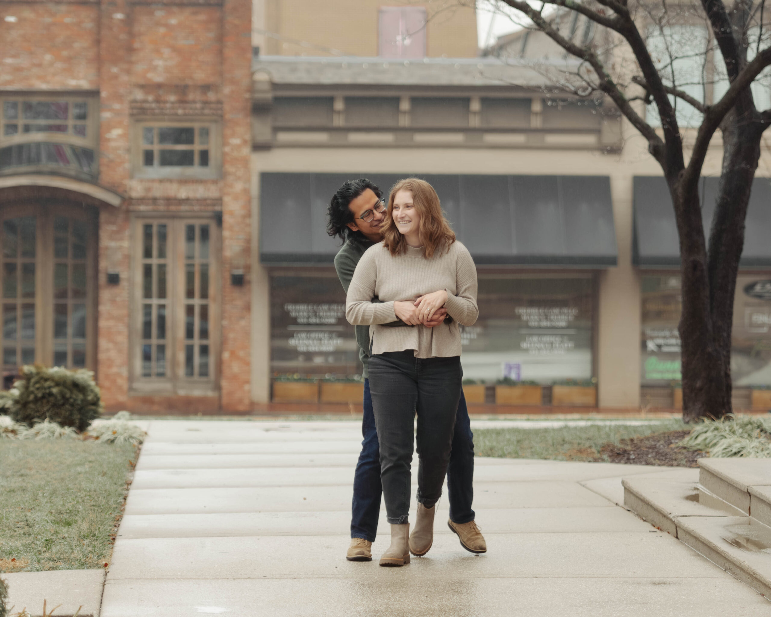 A man hugs a woman from behind during their engagement session, they are laughing 