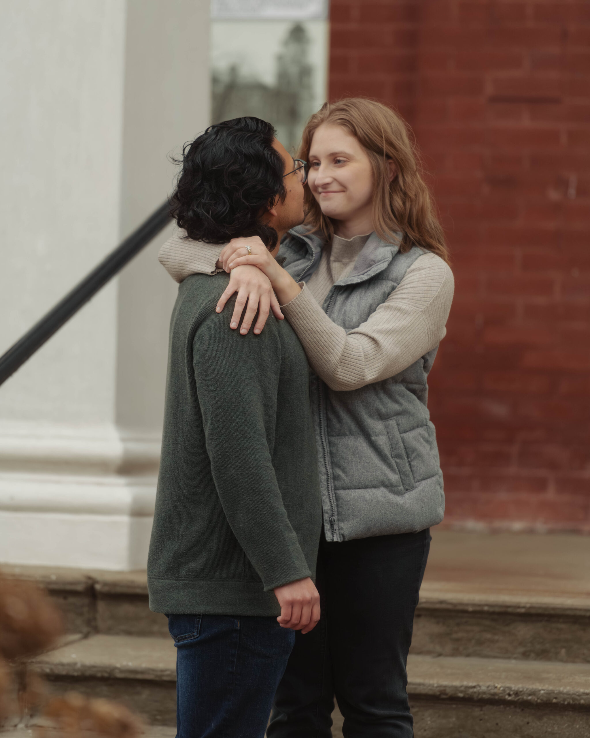 A man and woman stand on the steps of a courthouse during their engagement session. She is looking into his eyes and smiling.