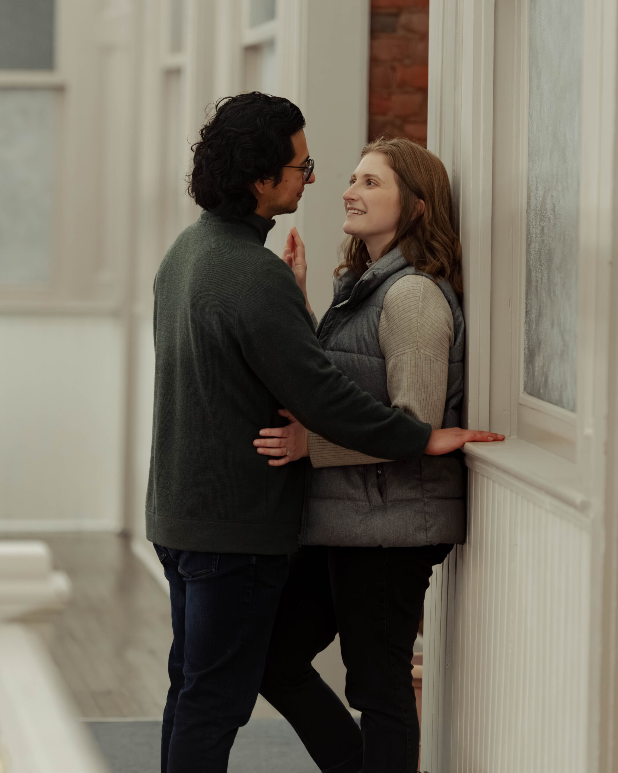 A woman and man face each other smiling and playing with each others hands. The woman's back is against the wall of an old building.