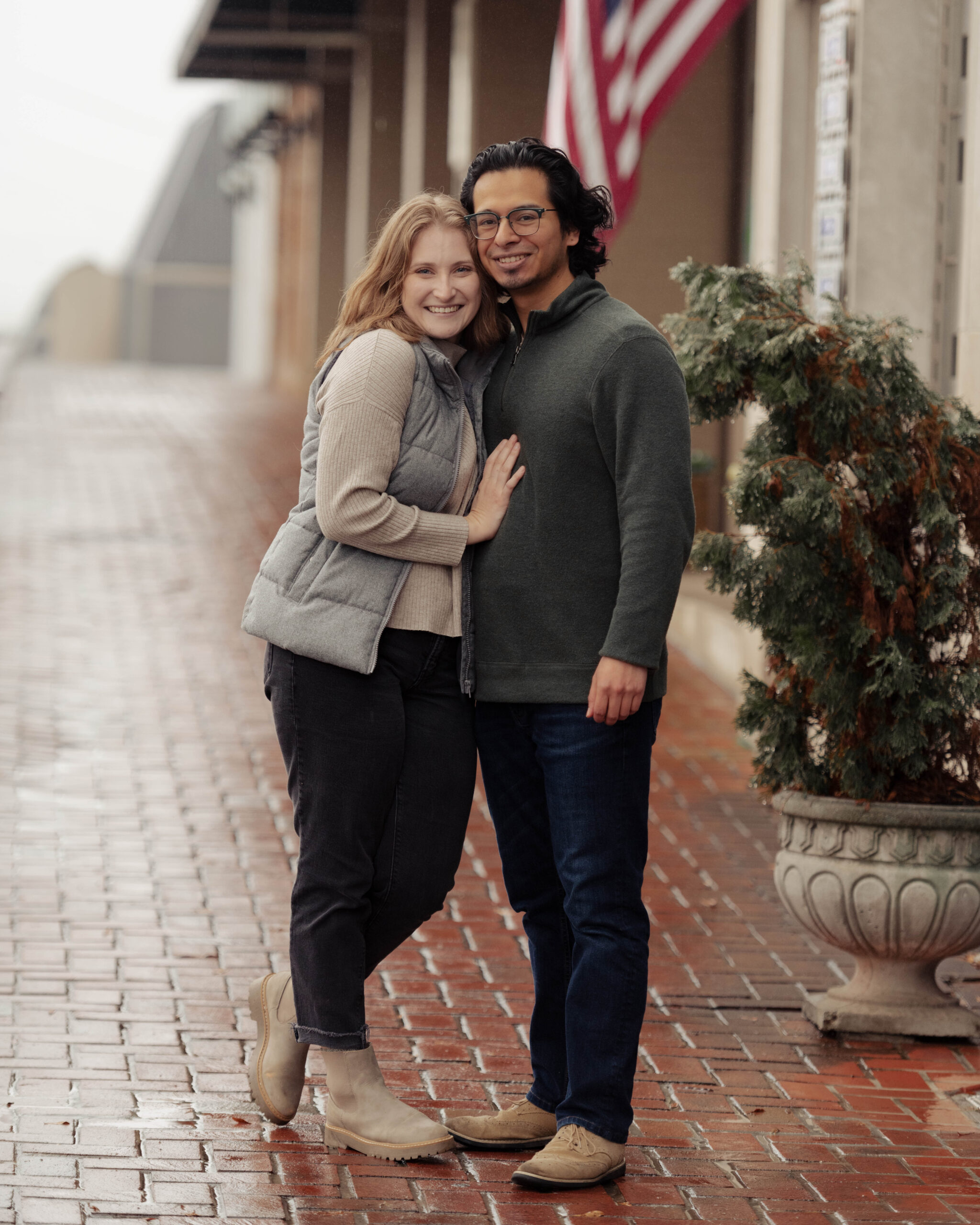 A white woman and Hispanic and/or Latino man look at the camera, hugging and smiling, on a rainy street.