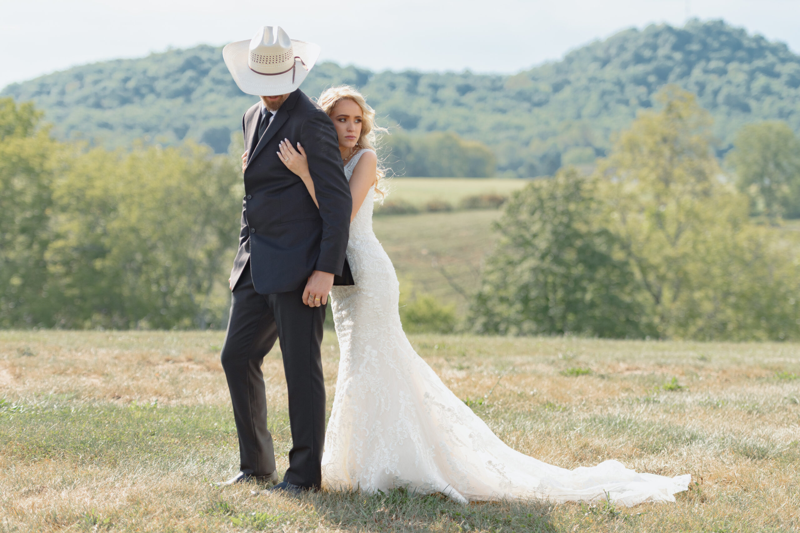 A bride and groom stand on a hill, the bride hugs the groom from behind. The groom is wearing a white cowboy hat and looking at the ground.