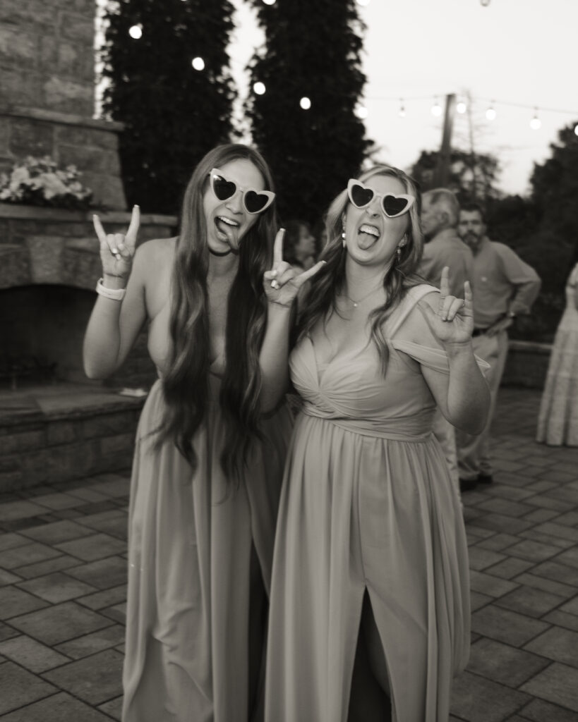 Two bridesmaids wearing heart-shaped sunglasses stick their tongues out at the camera.