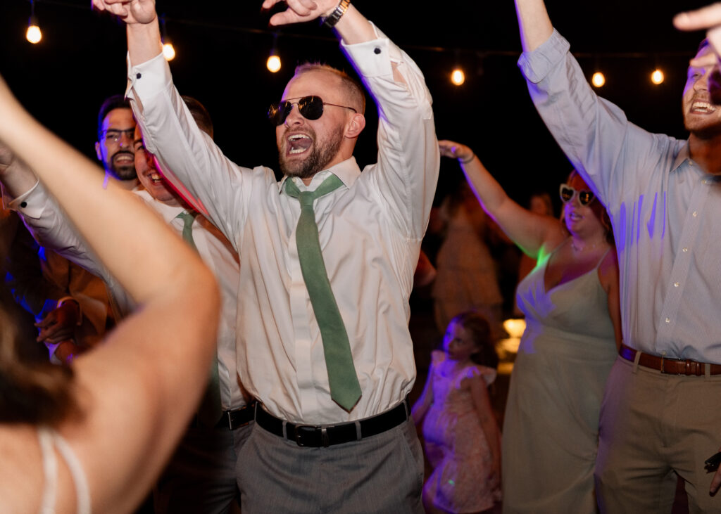 A groomsman dances at a wedding with sunglasses at night and his arms in the air.