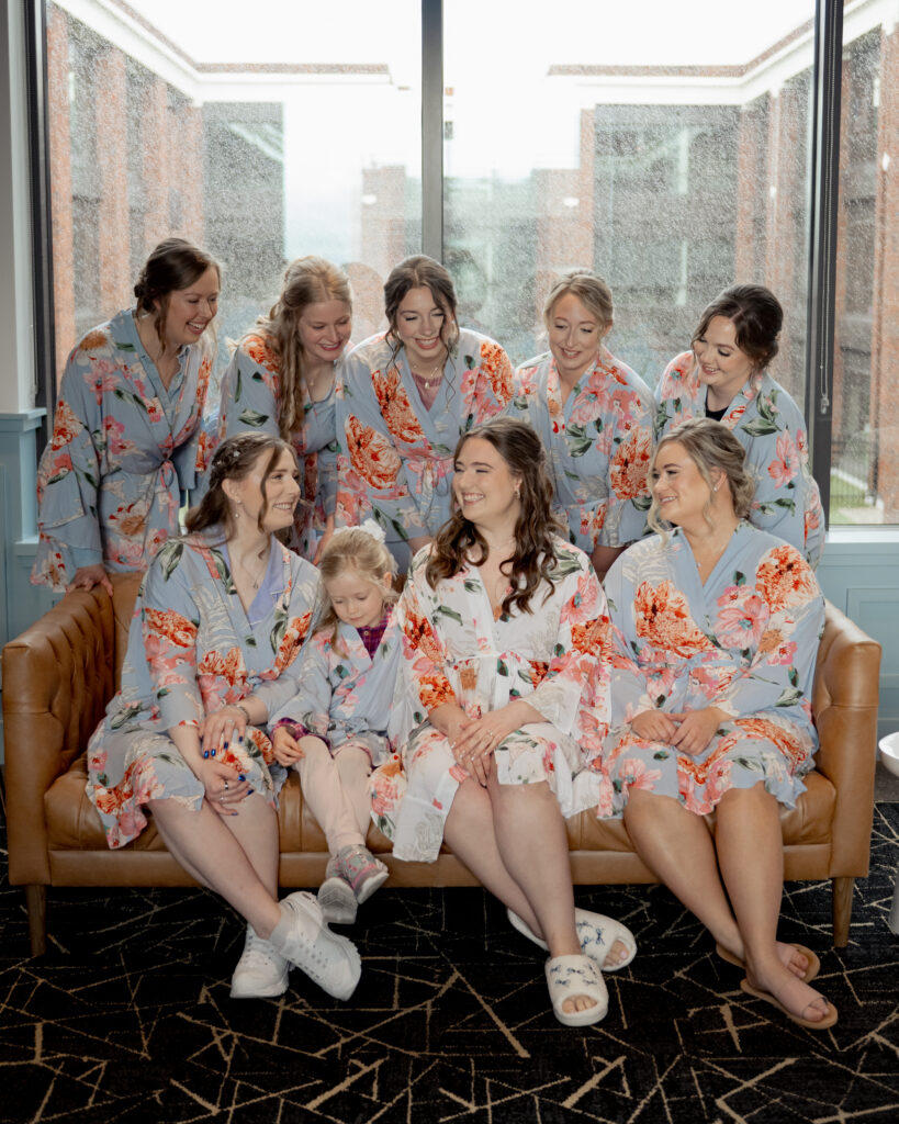 A group of bridesmaids gather around the bride as they all laugh in matching blue floral robes ahead of a catholic wedding ceremony.