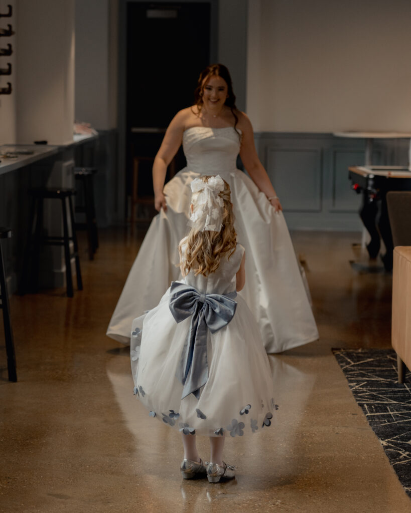 A Bride shows off her wedding dress to a flower girl wearing a dress with a big blue bow.