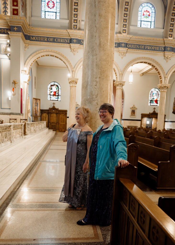 A bride's mom and mother in law watch her leave a bouquet of flowers for Mary before she gets married.