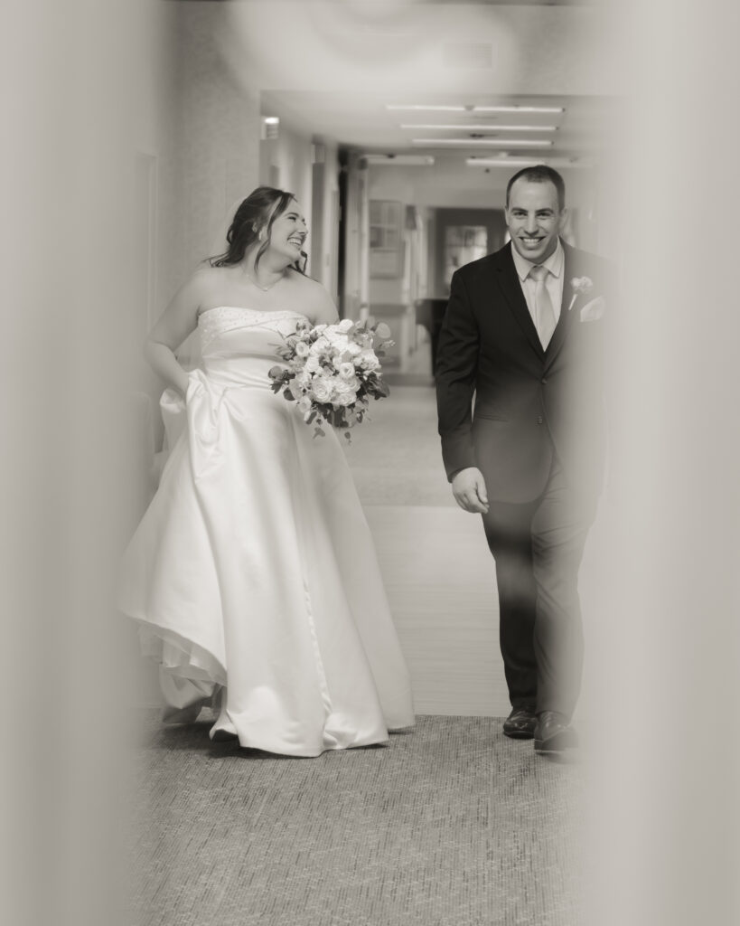 A catholic bride and groom walk down the hallway of a nursing home laughing after a wedding-day visit with the groom's grandmother.