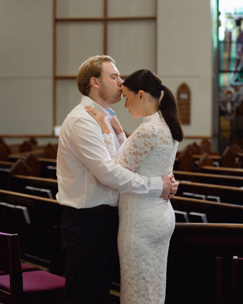 A couple poses inside St. Mary's Catholic church in Jackson, Tennessee during their sunny engagement session ahead of their catholic wedding. The man kisses the woman's forehead.