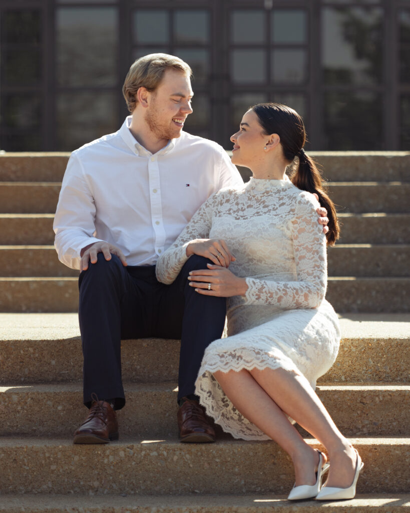 A catholic couple sits on the steps in front of St. Mary's Catholic church in Jackson, Tennessee during their sunny engagement session smiling at each other.