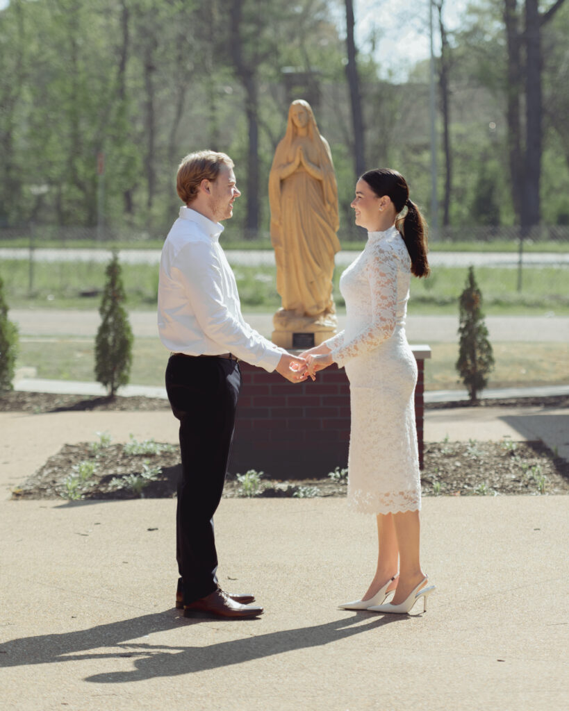 A catholic engaged couple pose in front of a statue of Mary at St. Mary's Catholic church in Jackson, Tennessee for their engagement session. They stand in front of the statue and hold hands in the direct sun.