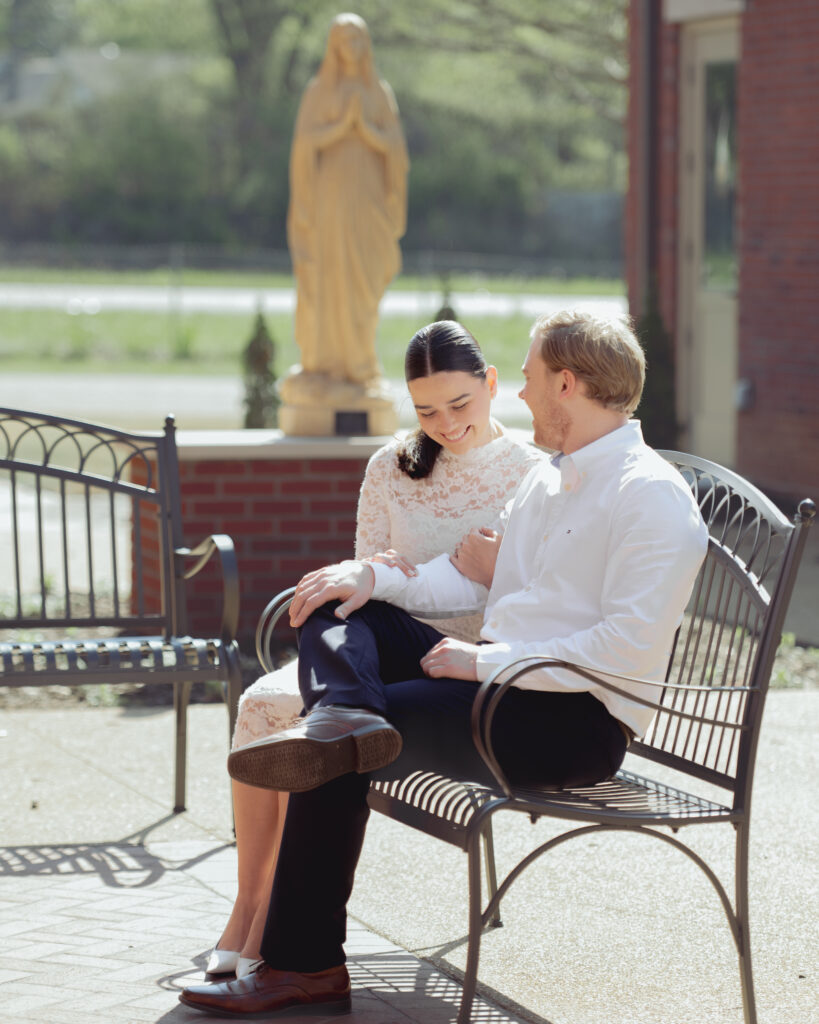 A catholic engaged couple pose in front of a statue of Mary at St. Mary's Catholic church in Jackson, Tennessee for their engagement session. They laugh together.