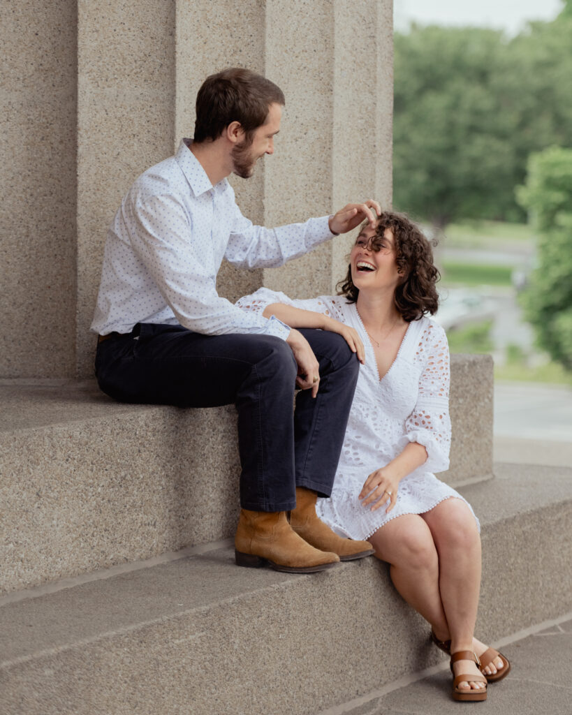 A catholic couple sits on the steps of the parthenon while the man brushes hair off the woman's forehead during their engagement at centennial park in nashville, tn
