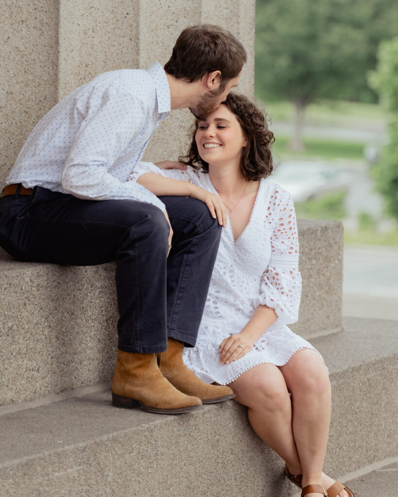 A catholic couple sits on the steps of the parthenon while the man gives the woman a kiss on the forehead during their engagement at centennial park in nashville, tn