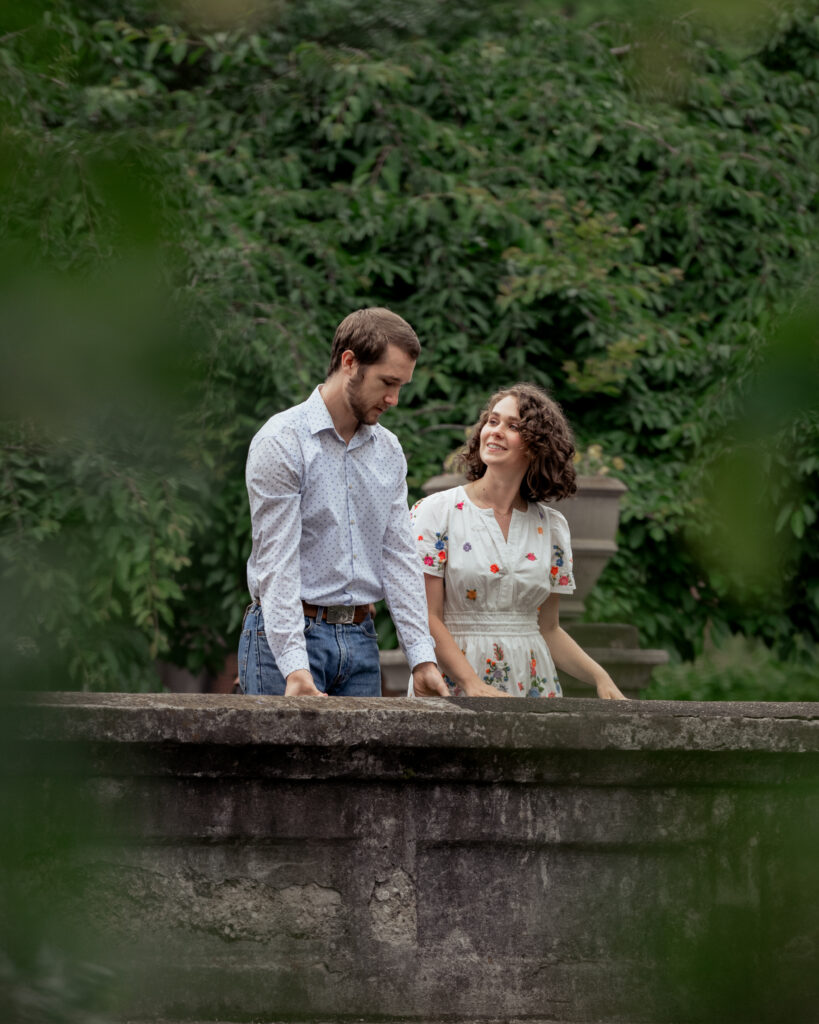 A catholic bride and groom have a conversation on a bridge framed by leaves in the foreground during their catholic engagement session at centennial park in nashville, tn