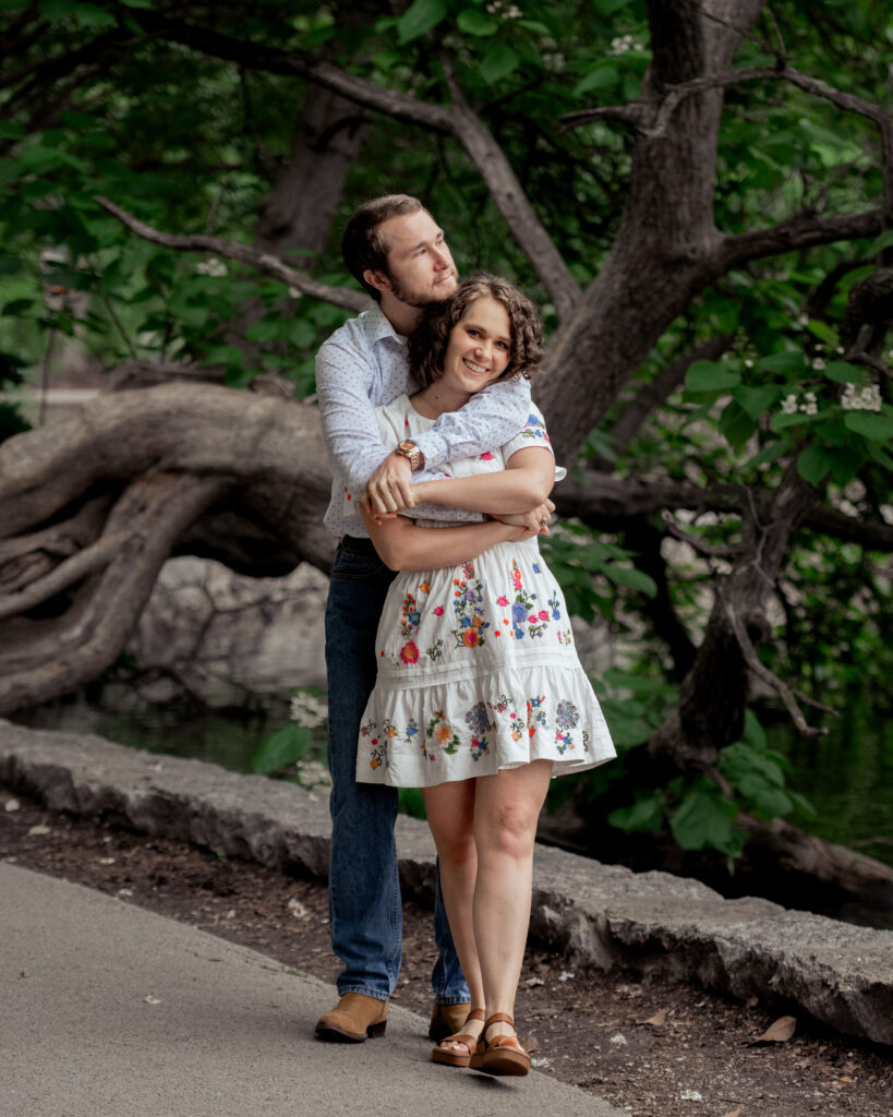 A catholic groom hugs a catholic bride from behind in front of a twisty tree during their catholic engagement session at centennial park in nashville, tn