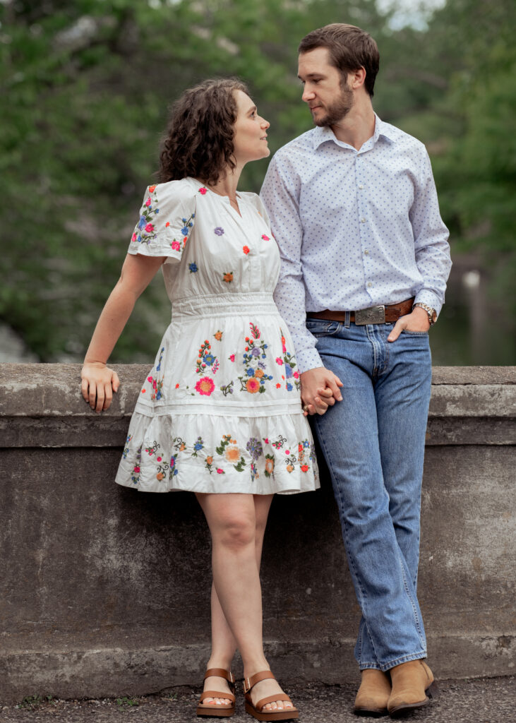 A catholic bride and groom lean against the side of a bridge in a relaxed pose and stare at each other during their catholic engagement session at centennial park in nashville, tn