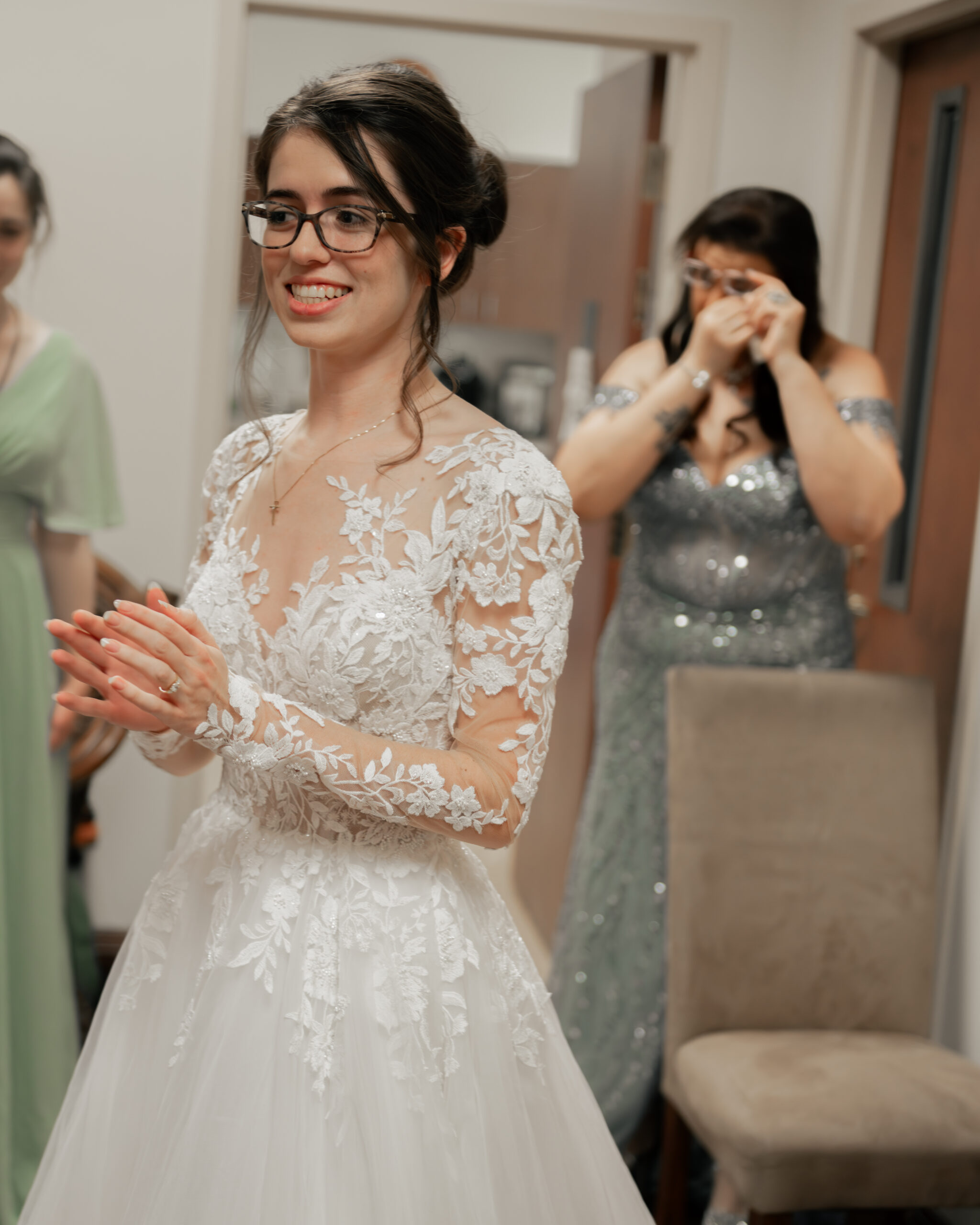 A bride smiles joyfully as her mother in law wipes tears away in the backround at Holy Family catholic church in Nashville ahead of a catholic wedding Mass.
