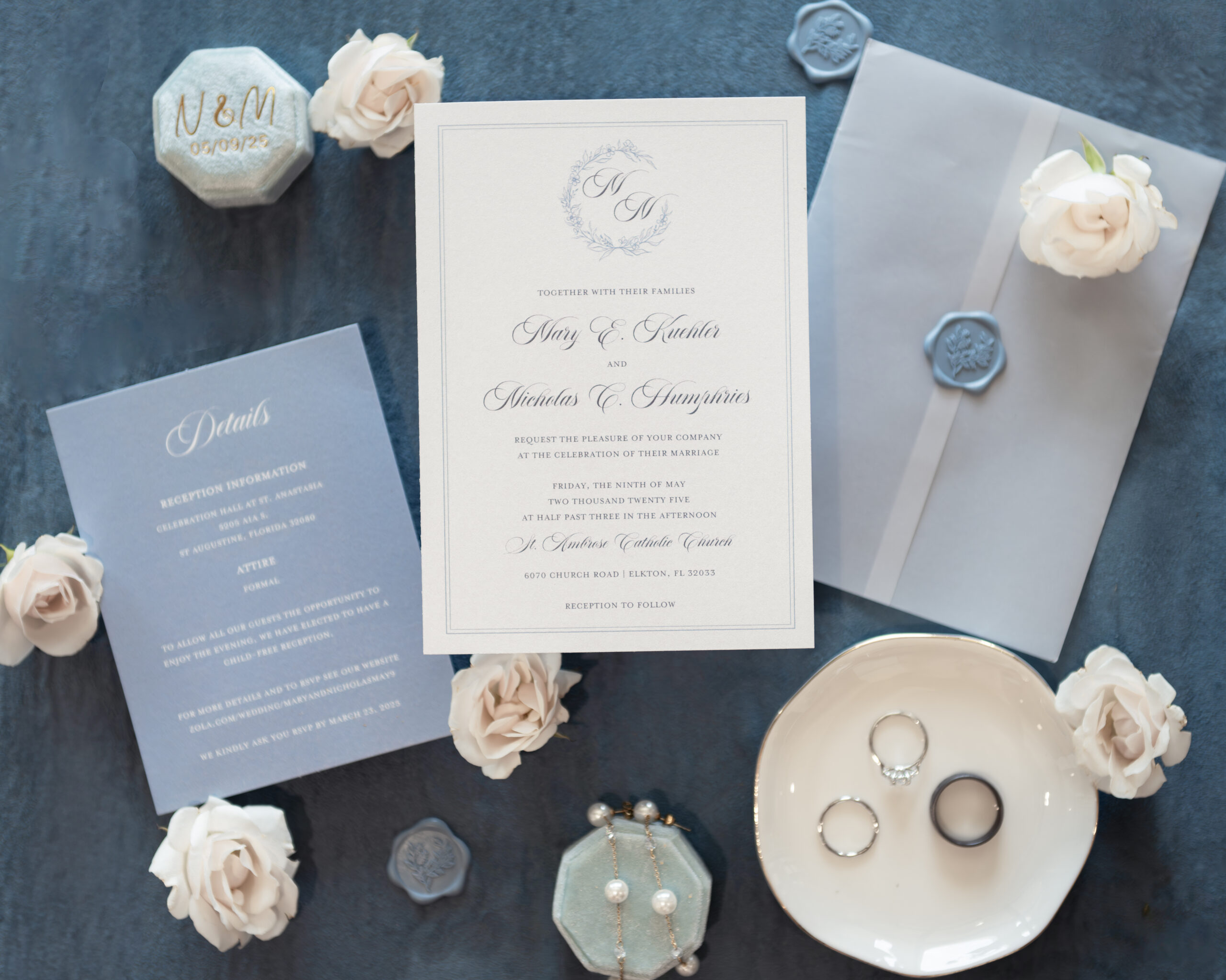 A flat lay photo of a Catholic wedding invitation, featuring a beautifully designed card with the couple's names, reception details, and wedding rings. Soft white roses and wax seals accentuate the elegant wedding stationery.