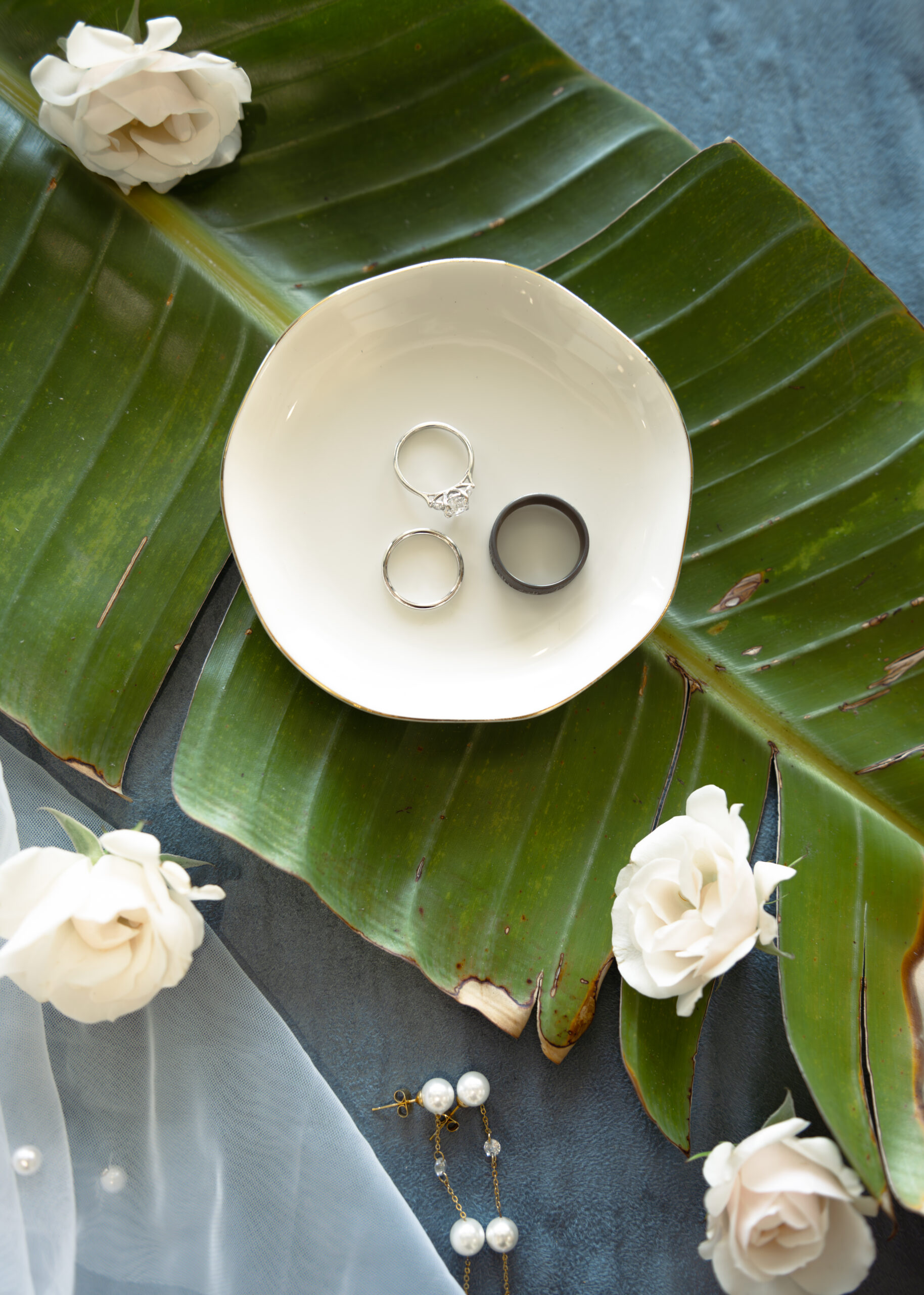 A close-up of a bride's wedding rings displayed on a plate, surrounded by white roses and green tropical leaves. The photo captures the timeless beauty of the wedding details during the getting ready portion of a Catholic wedding