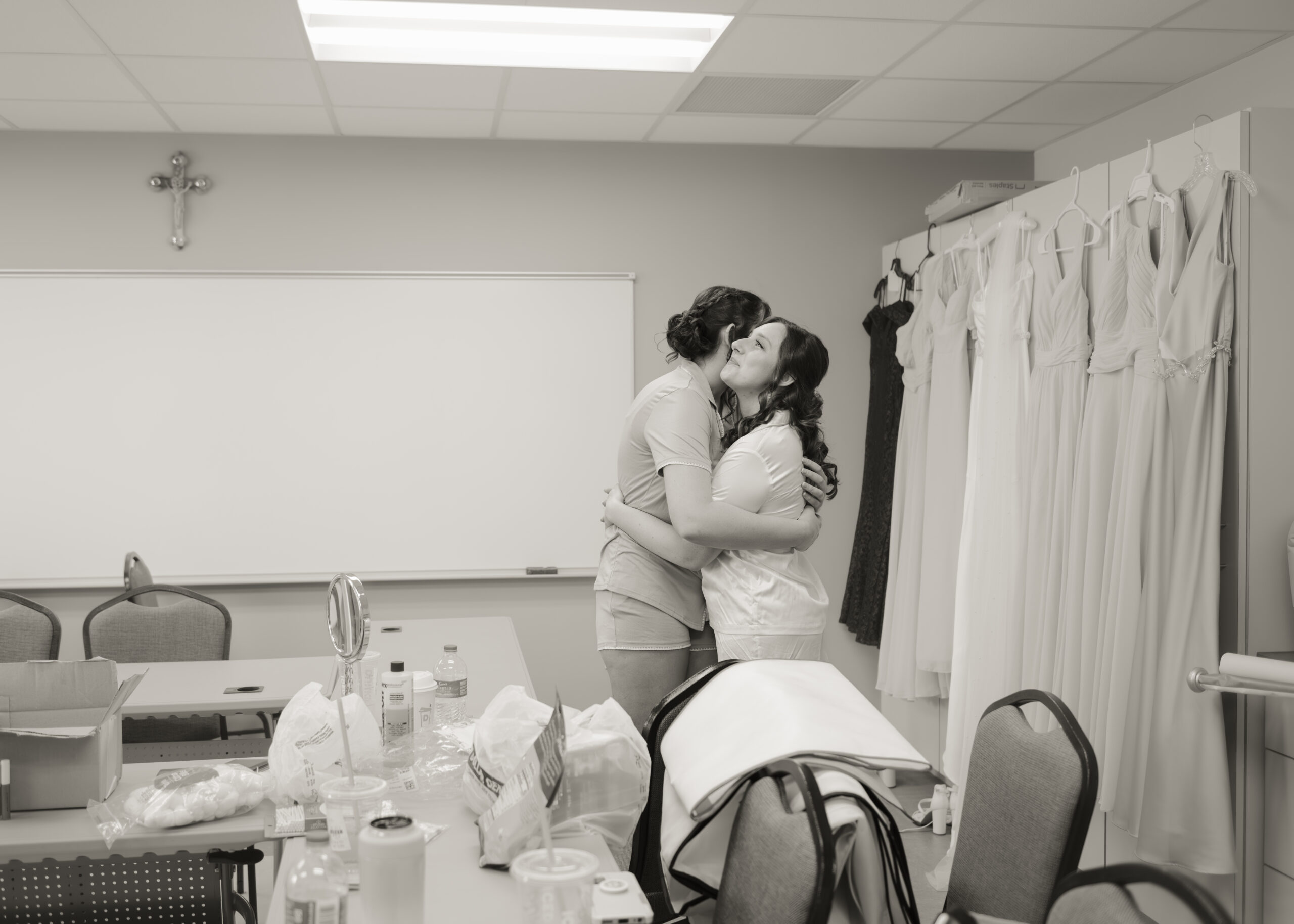 A black-and-white photo capturing an emotional moment between the bride and her sister, hugging and sharing a joyful moment while preparing for the wedding. The photo captures the love and support shared between the bridal party before the Catholic wedding ceremony.