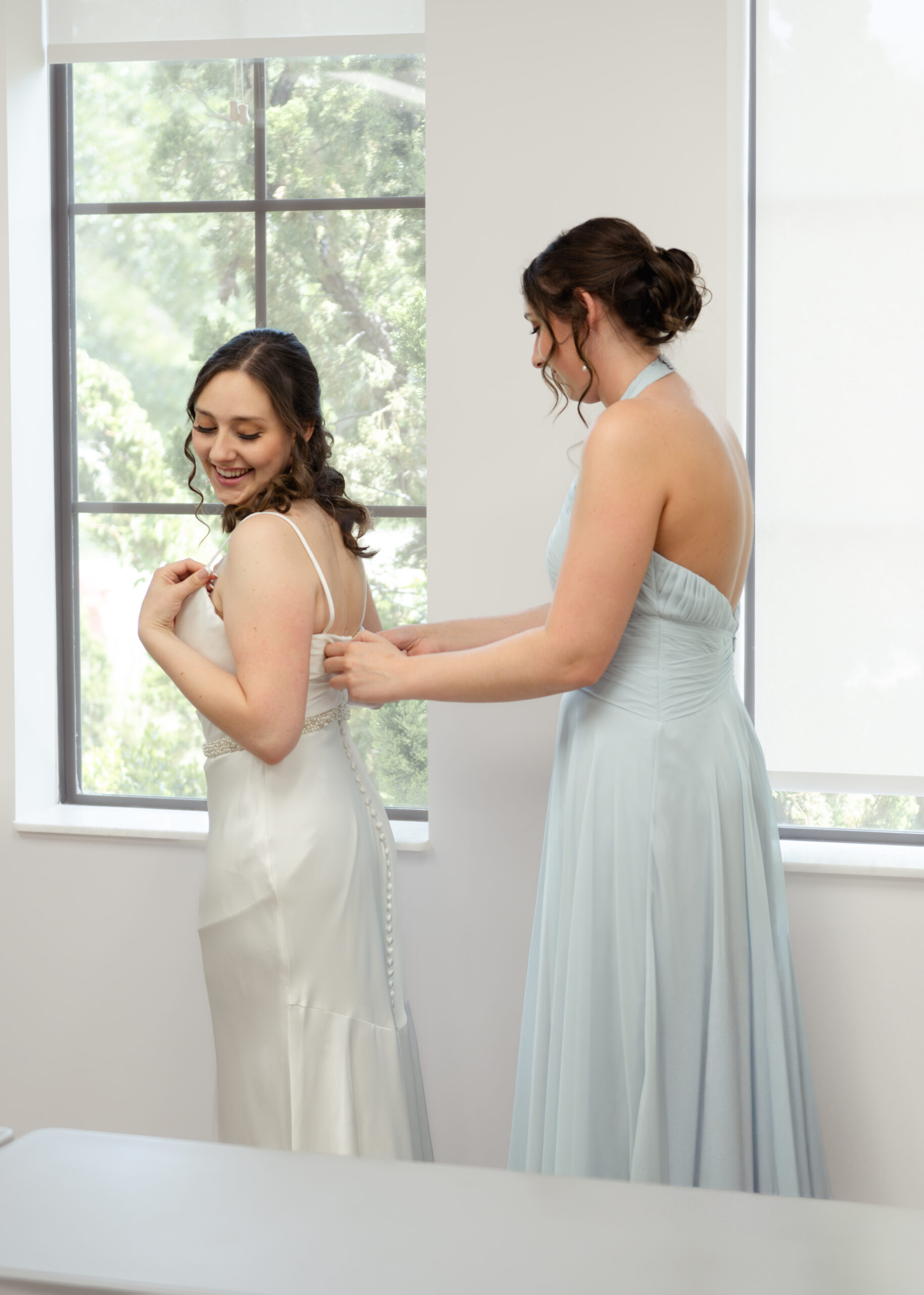 A bridesmaid helps the bride with the final touches of her wedding dress during the getting ready moments before the Catholic wedding ceremony. The bride smiles happily, surrounded by natural light from the window, showcasing the beauty of the day.
