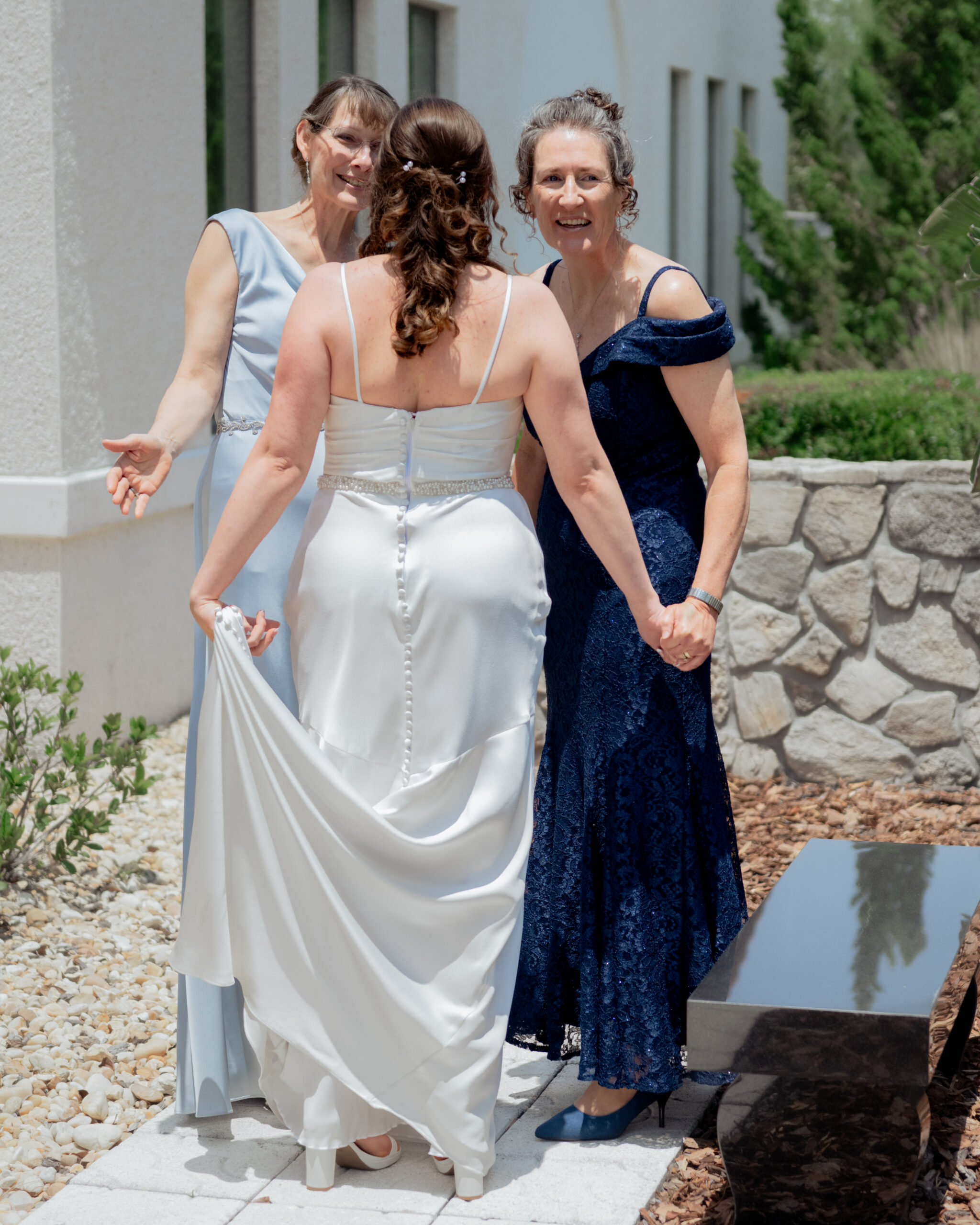 A heartfelt moment during a Catholic wedding in St. Augustine, Florida, as the bride shares a first look with her mother and mother-in-law. The bride in her satin gown stands with both women, all holding hands and smiling as they admire her stunning dress before the ceremony.