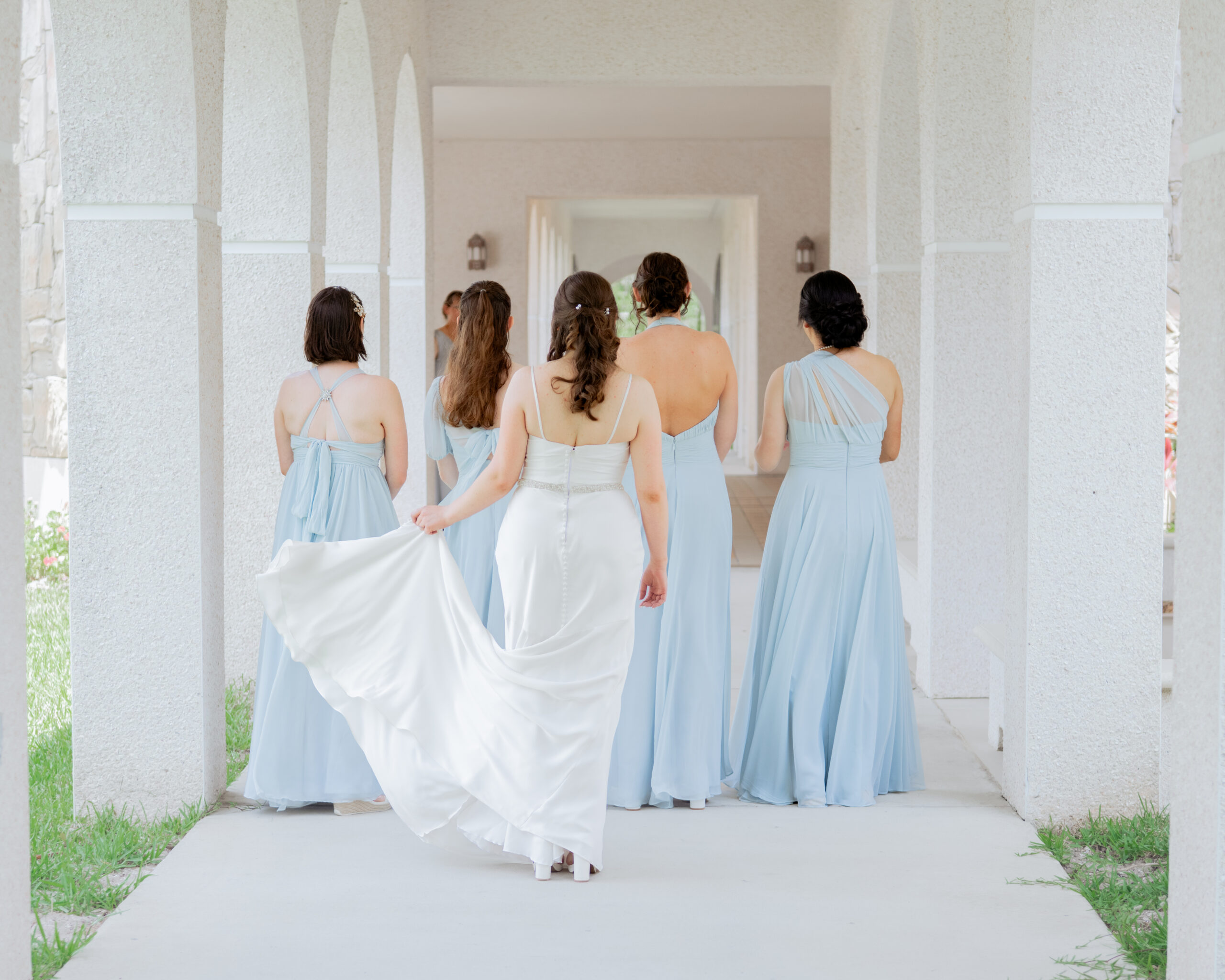 The bride and her bridesmaids share an emotional first look before the Catholic wedding ceremony in St. Augustine, Florida. The bridesmaids, dressed in elegant blue gowns, stand together as they marvel at the bride in her satin wedding dress, capturing a joyful moment of support and excitement.