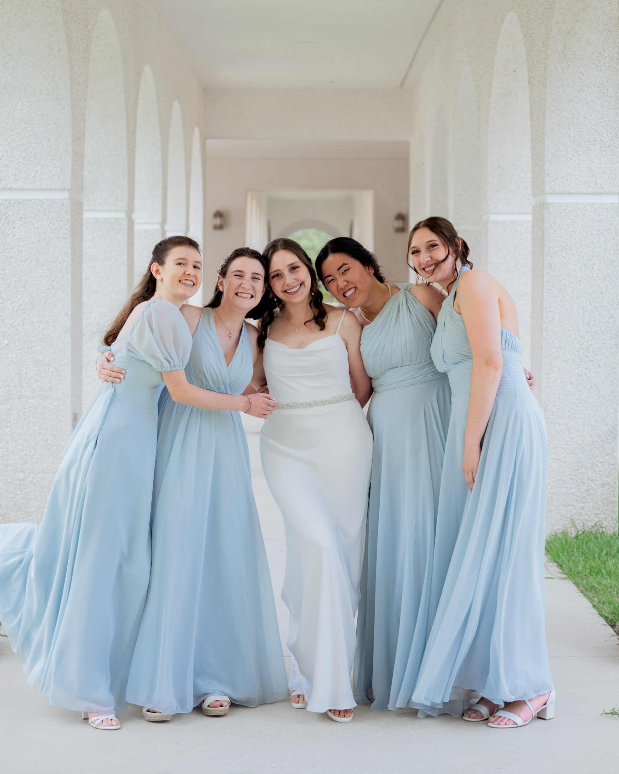 A tender moment as the bride, wearing a satin gown, stands surrounded by her bridesmaids in blue dresses. They embrace and smile together, celebrating the bride’s first look before the Catholic wedding in St. Augustine, Florida. This image captures the love and excitement of the bride’s closest friends.