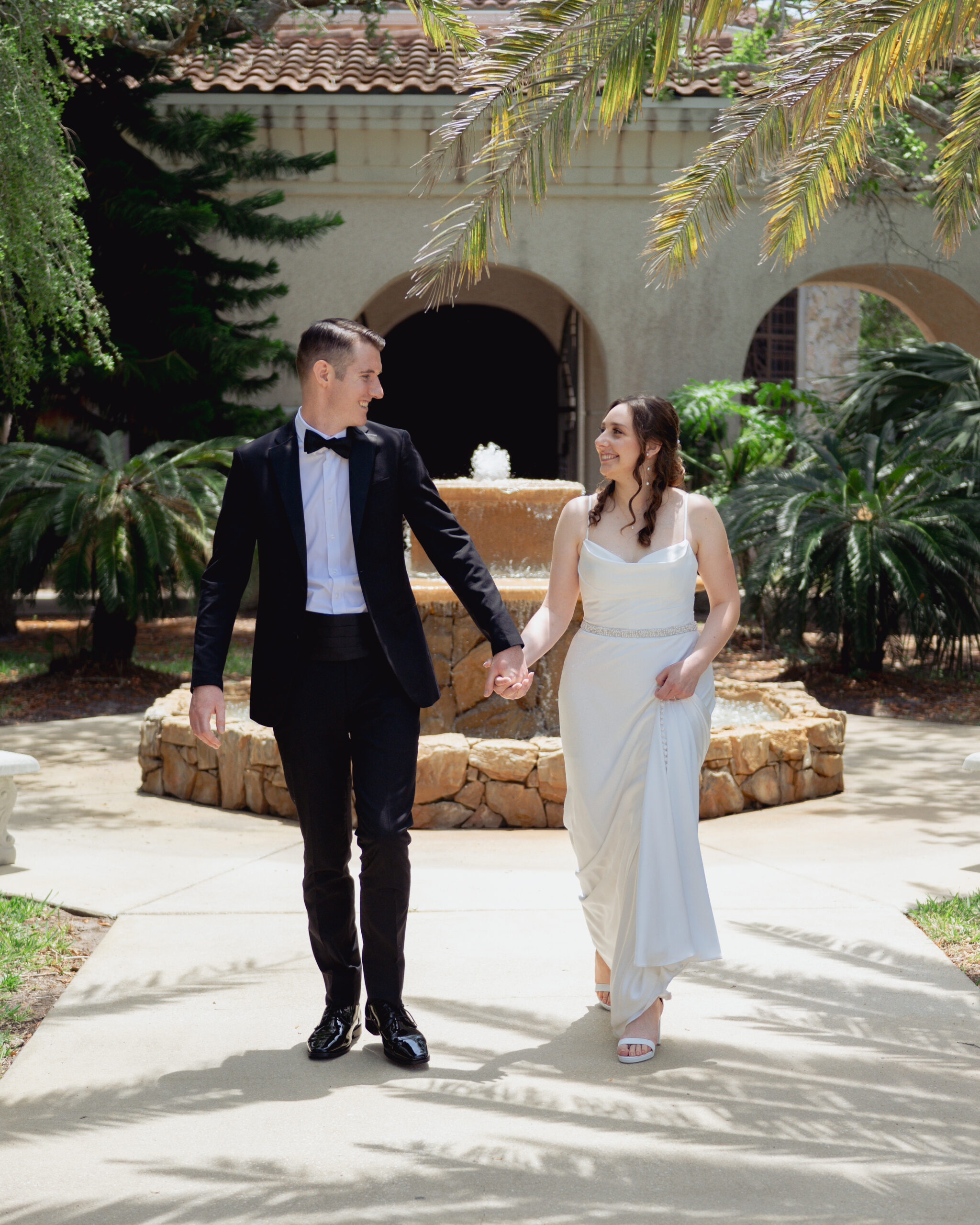 Catholic bride and groom holding hands and walking joyfully outside St. Anastasia’s Catholic Church before their wedding mass, surrounded by tropical greenery and a stone fountain.