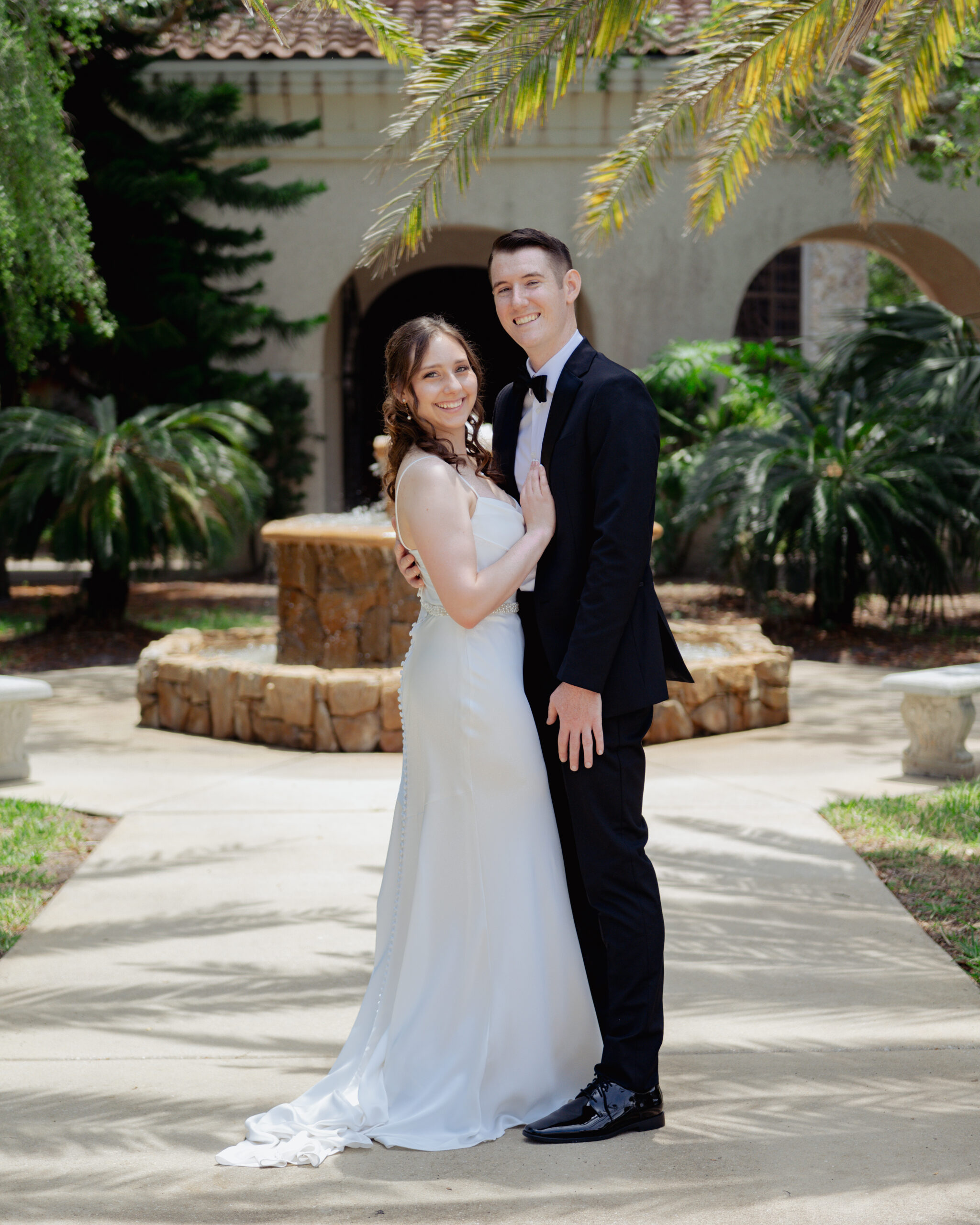 Bride and groom posing in front of the fountain at St. Anastasia’s Catholic Church, smiling and embracing before their Catholic wedding ceremony.