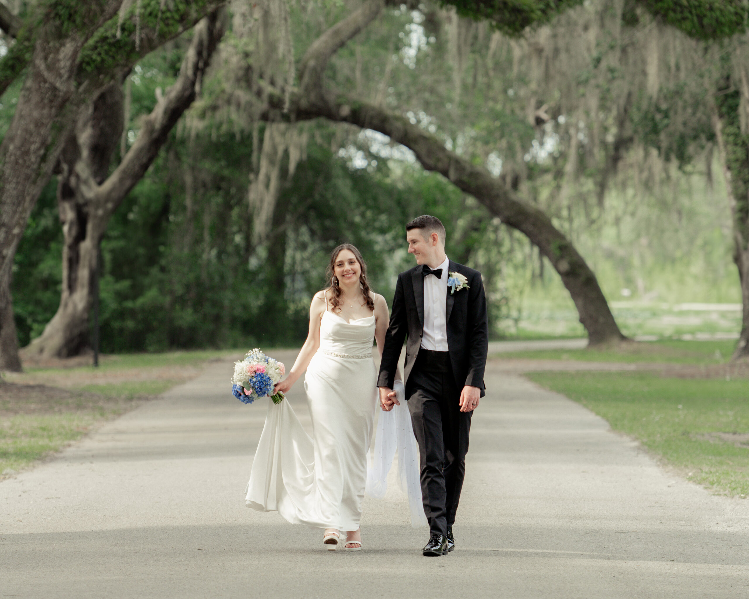 Bride and groom walking hand in hand under moss-draped oak trees at St. Ambrose Catholic Church in Elkton, Florida, during a romantic wedding portrait session.