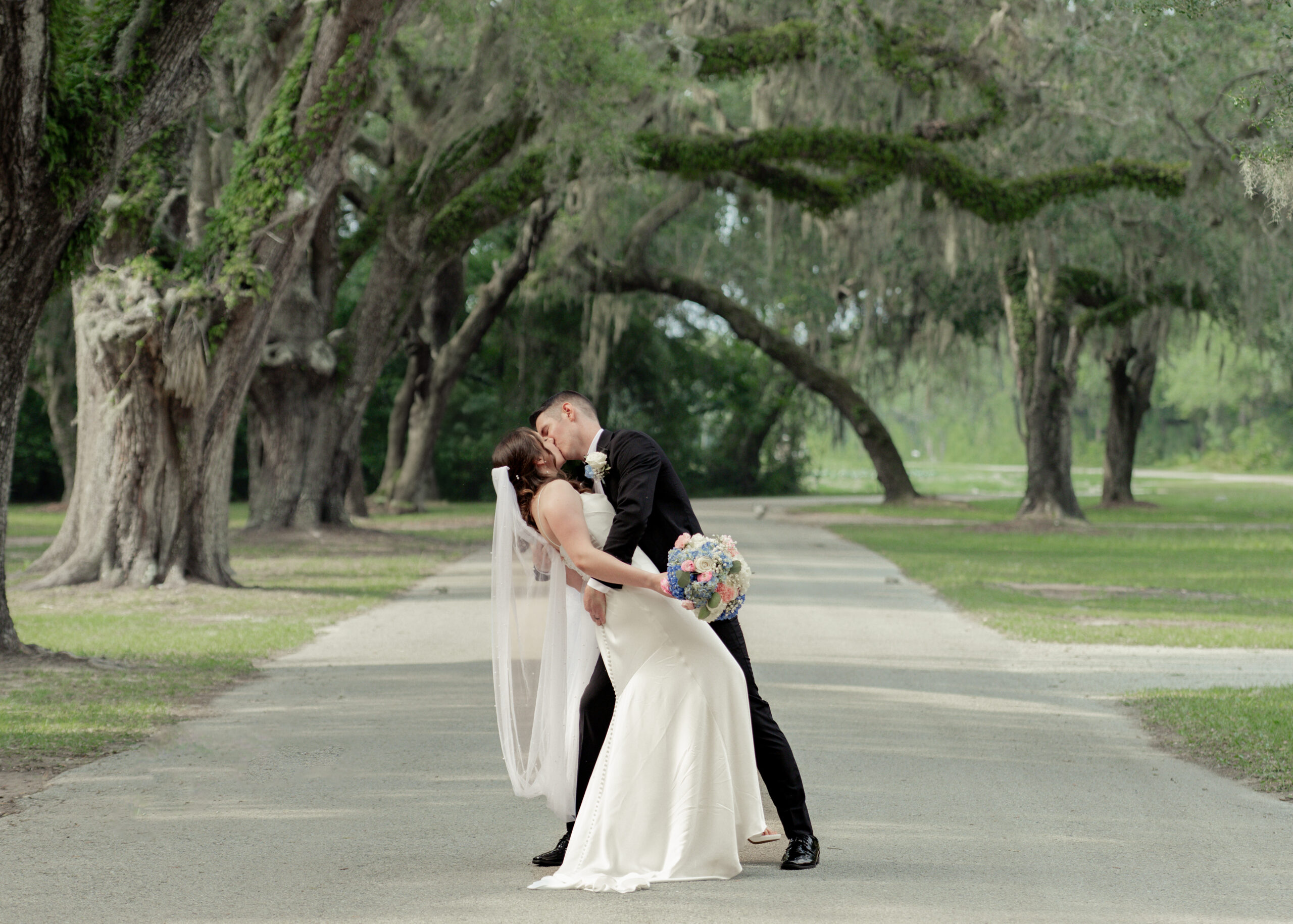 Groom dips and kisses bride on a tree-lined path at St. Ambrose Catholic Church in Elkton, Florida, capturing a timeless and elegant wedding moment.
