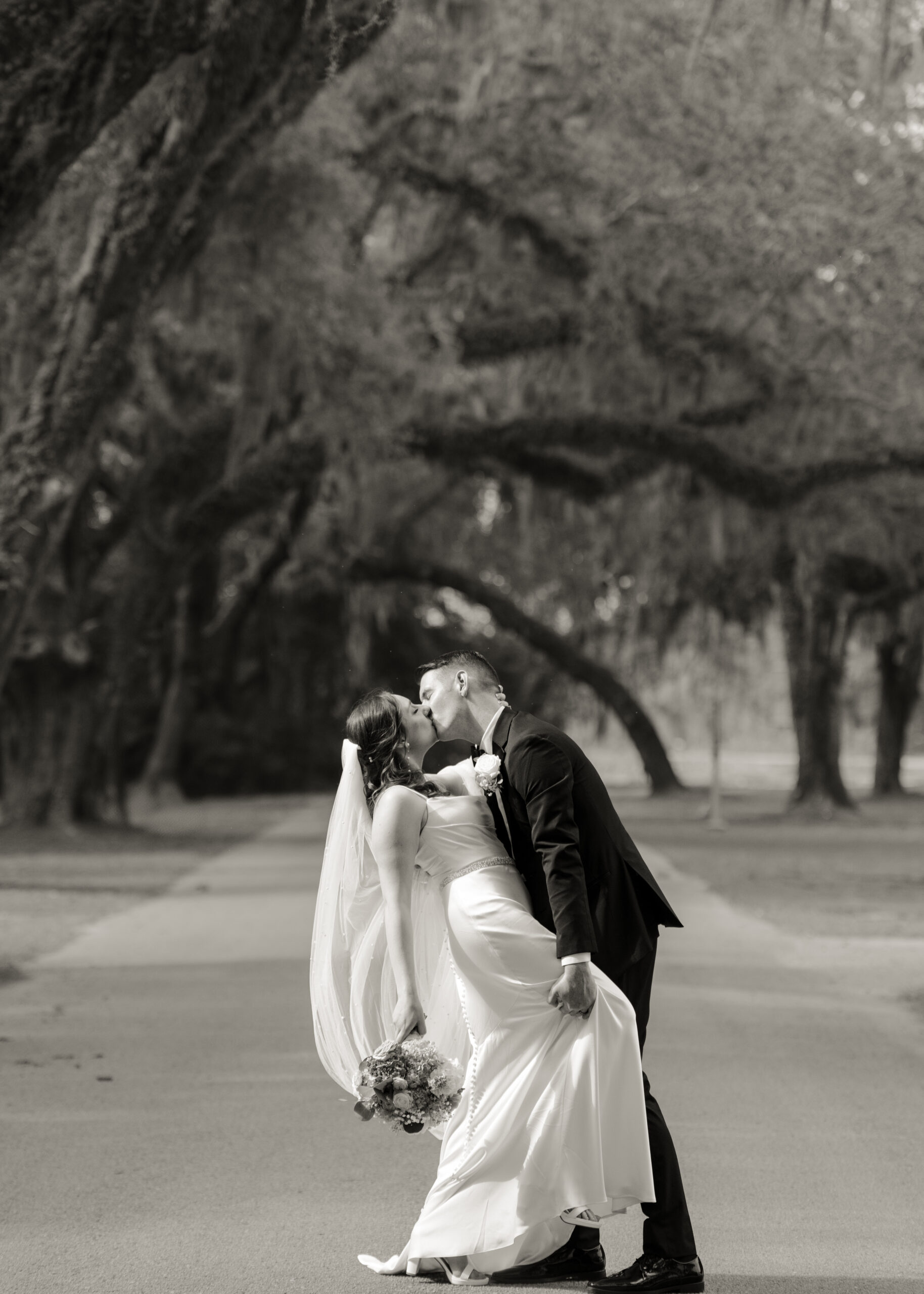 Black and white bridal portrait of groom dipping and kissing bride beneath the majestic oak canopy at St. Ambrose Catholic Church in Elkton, Florida.