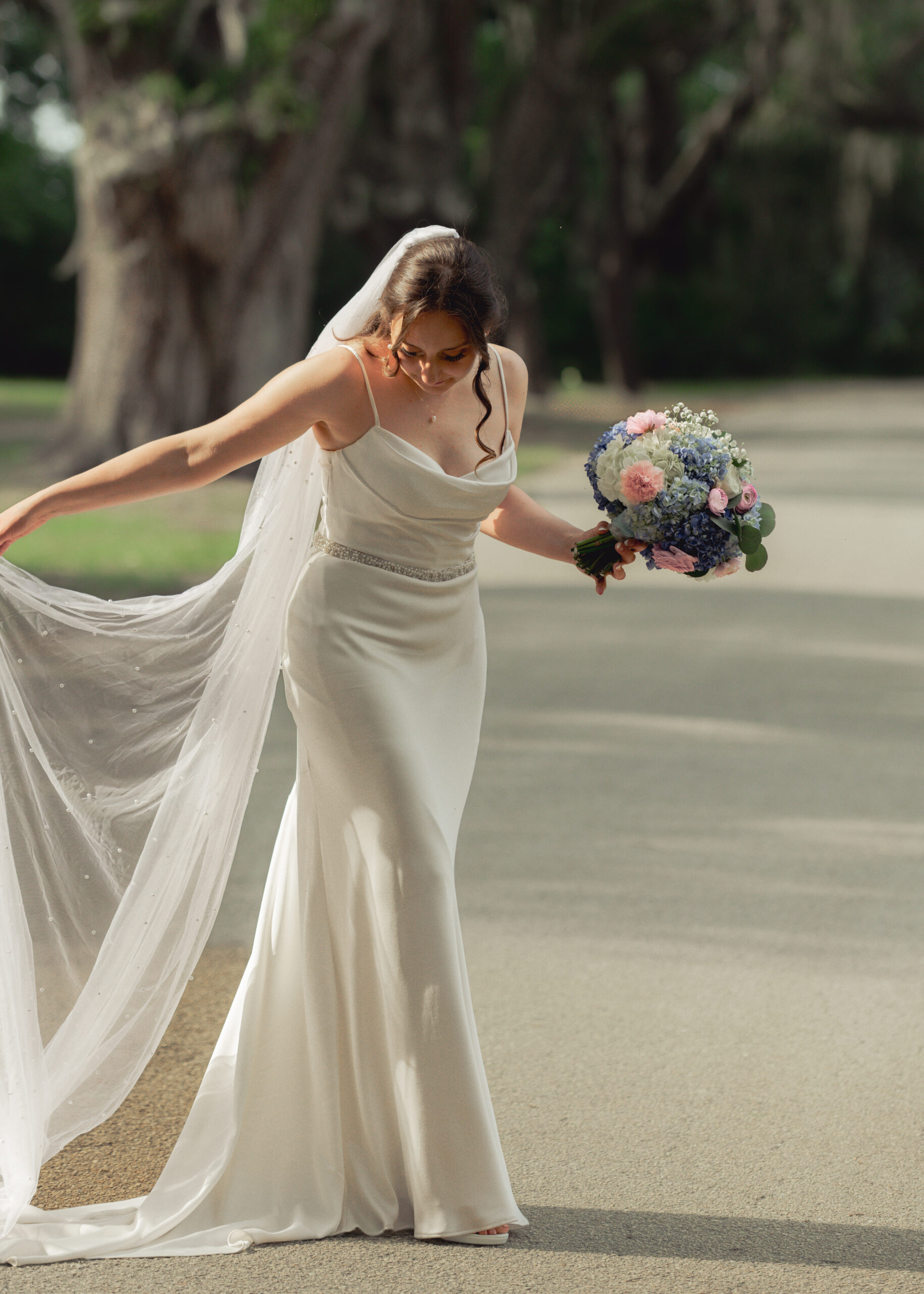 Elegant bride holding blue and pink bouquet and lifting her dress gracefully on the church pathway at St. Ambrose Catholic Church in Elkton, Florida.