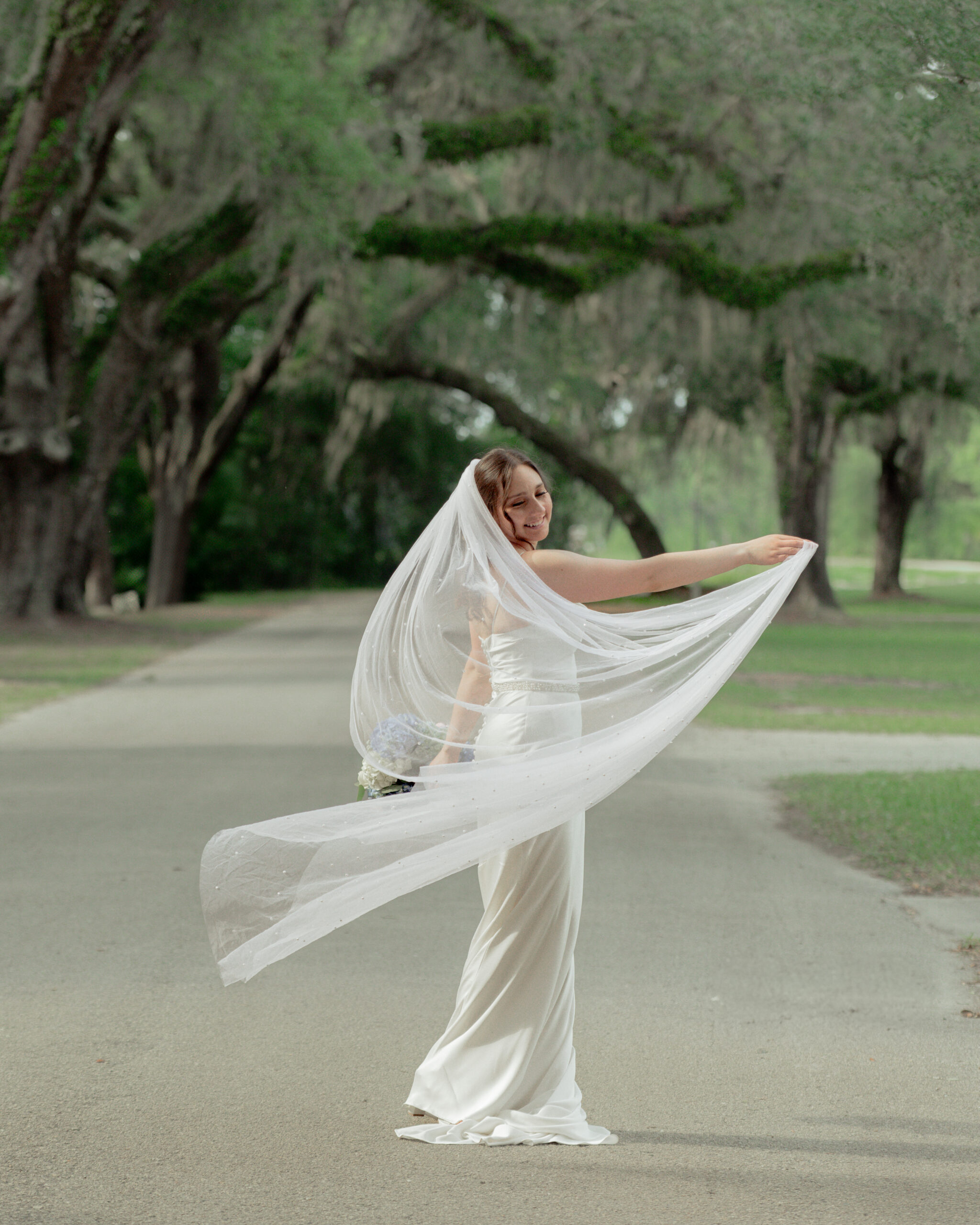 Joyful bride twirling her long flowing veil on the scenic tree-lined road near St. Ambrose Catholic Church in Elkton, Florida, during bridal portraits.