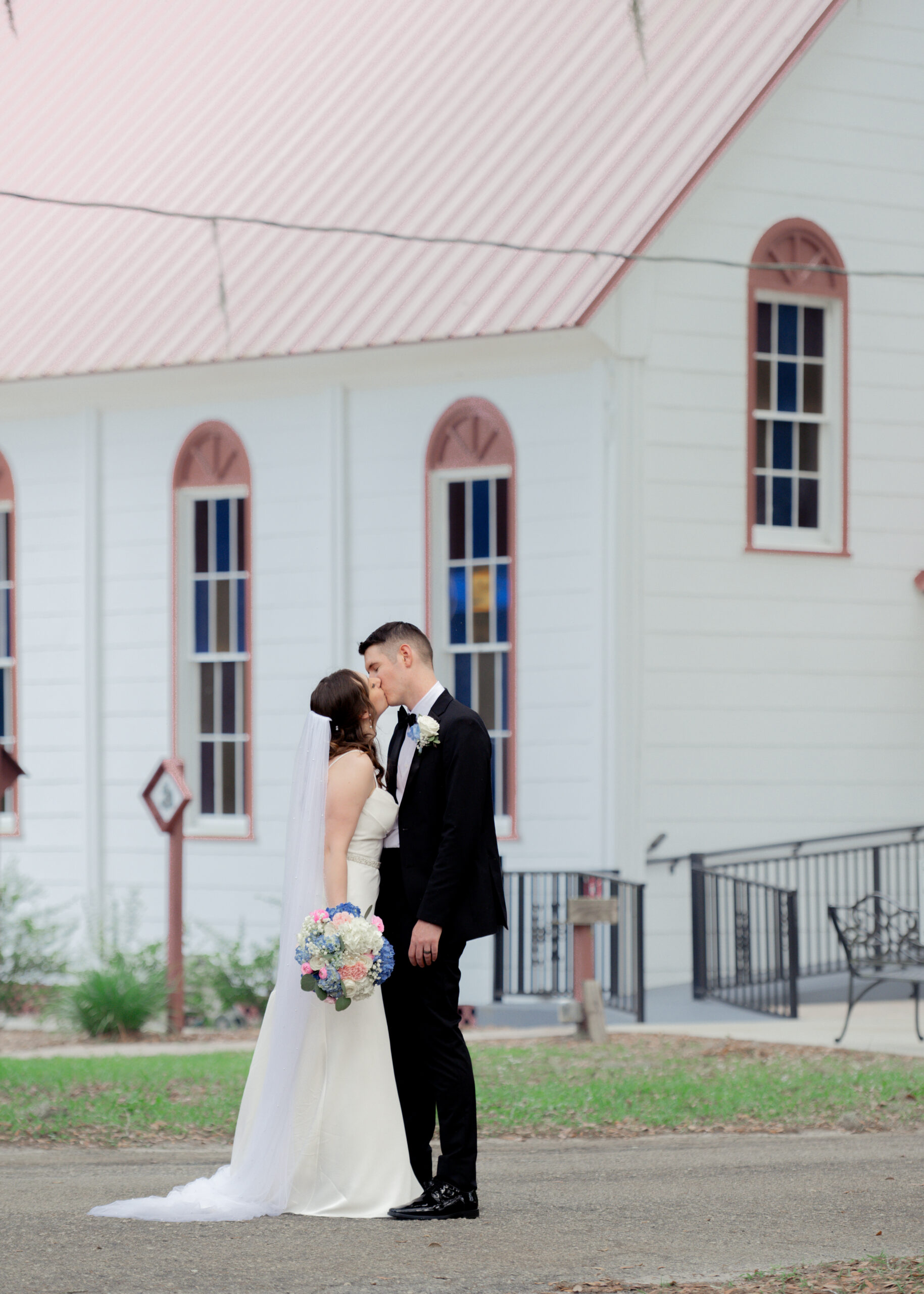 Bride and groom sharing a kiss in front of the historic white exterior of St. Ambrose Catholic Church in Elkton, Florida, after their wedding ceremony.
