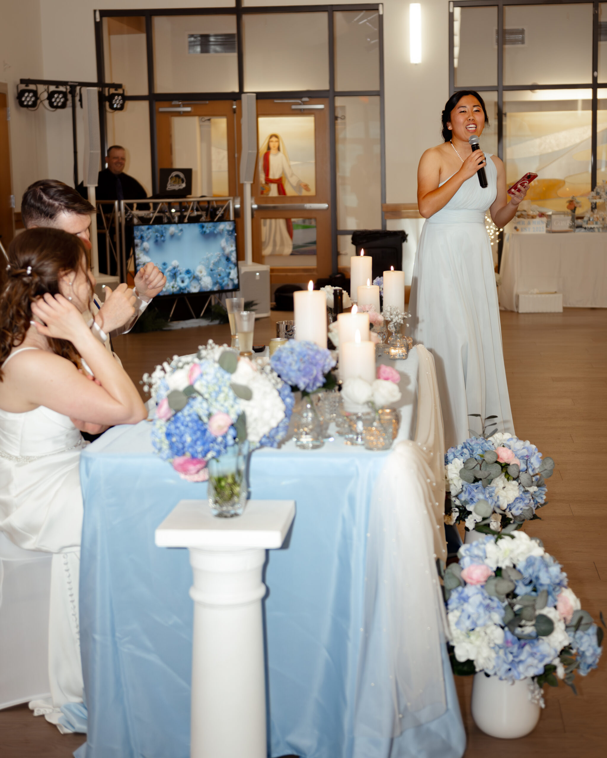 Maid of honor gives an emotional wedding speech with a microphone at a St. Augustine Catholic wedding reception at St. Anastasia Church. Elegant decor with candles and blue floral arrangements sets a romantic ambiance.