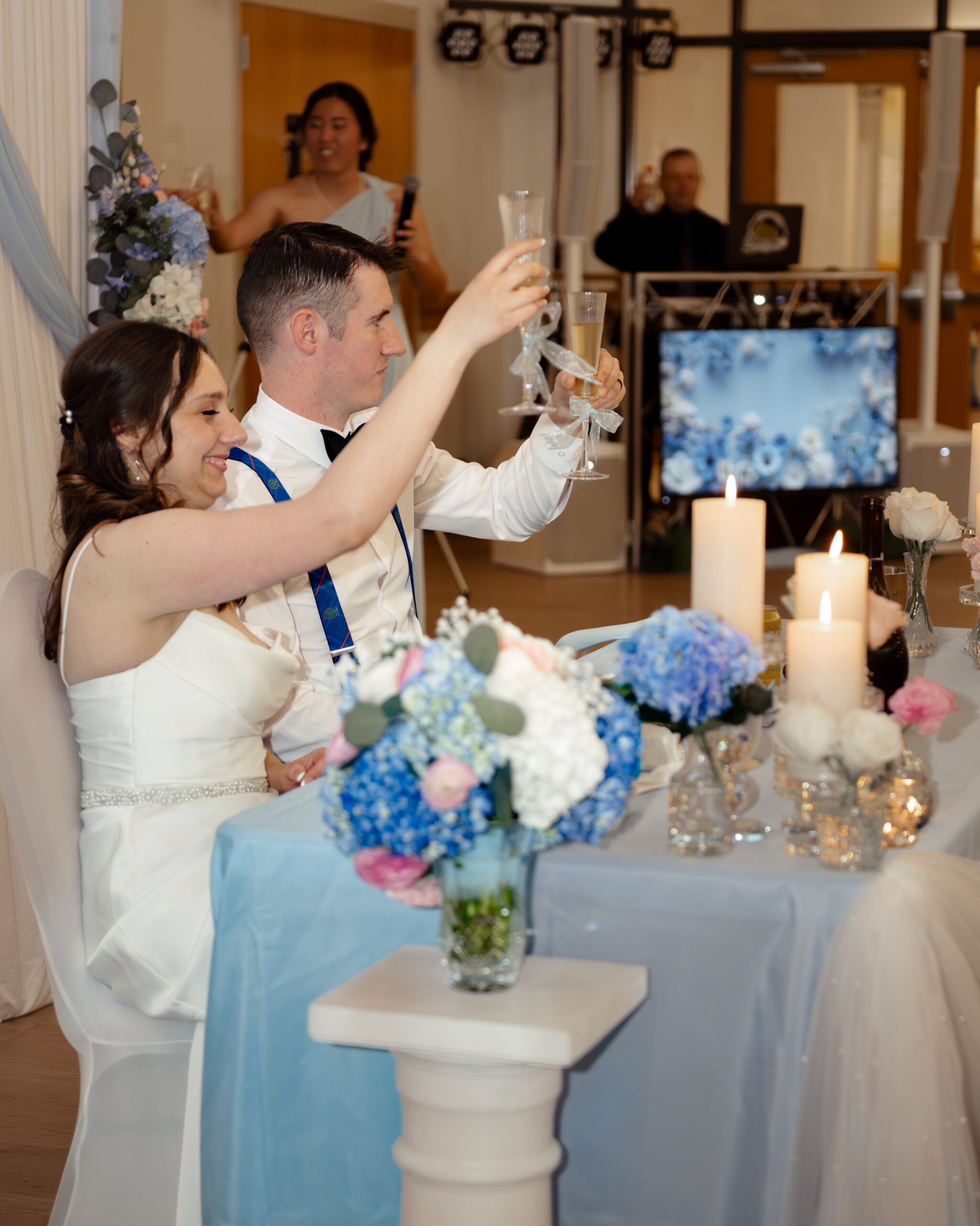 ride and groom raise champagne glasses during a toast as the maid of honor speaks in the background at their Catholic wedding reception at St. Anastasia in St. Augustine, Florida. The setting features soft blue tones and romantic floral decor.
