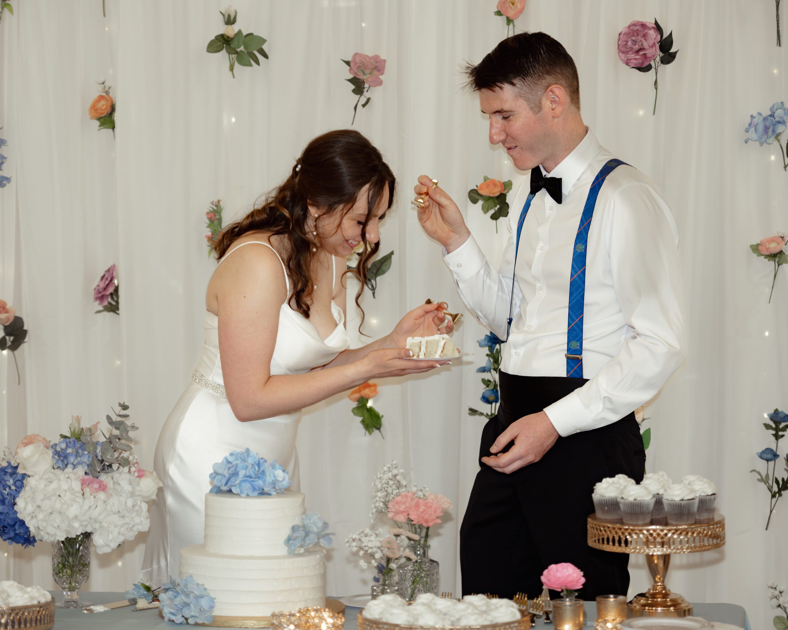 Bride and groom share a joyful moment cutting the wedding cake at their St. Augustine reception at St. Anastasia Catholic Church. The cake table is decorated with fresh flowers and elegant desserts.