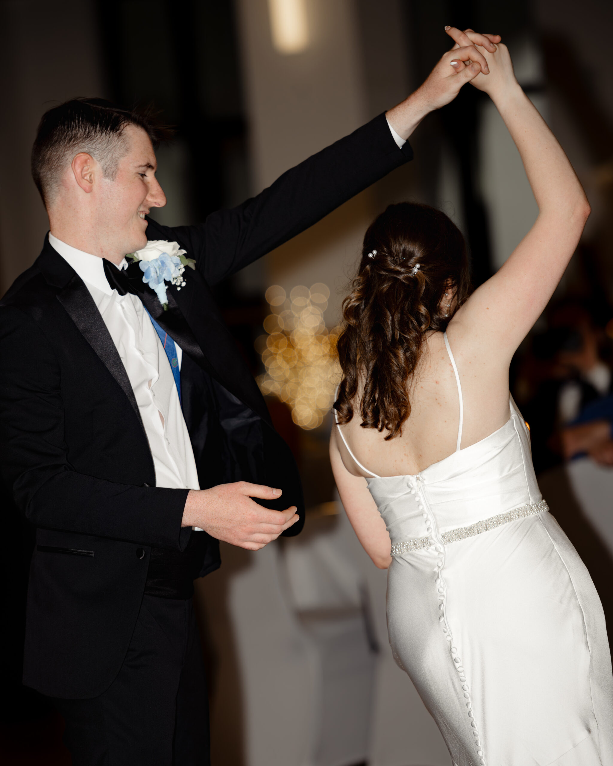 Groom twirls the bride during their first dance at a Catholic wedding reception at St. Anastasia Catholic Church in St. Augustine, Florida. Her satin wedding dress sparkles under the soft reception lighting.