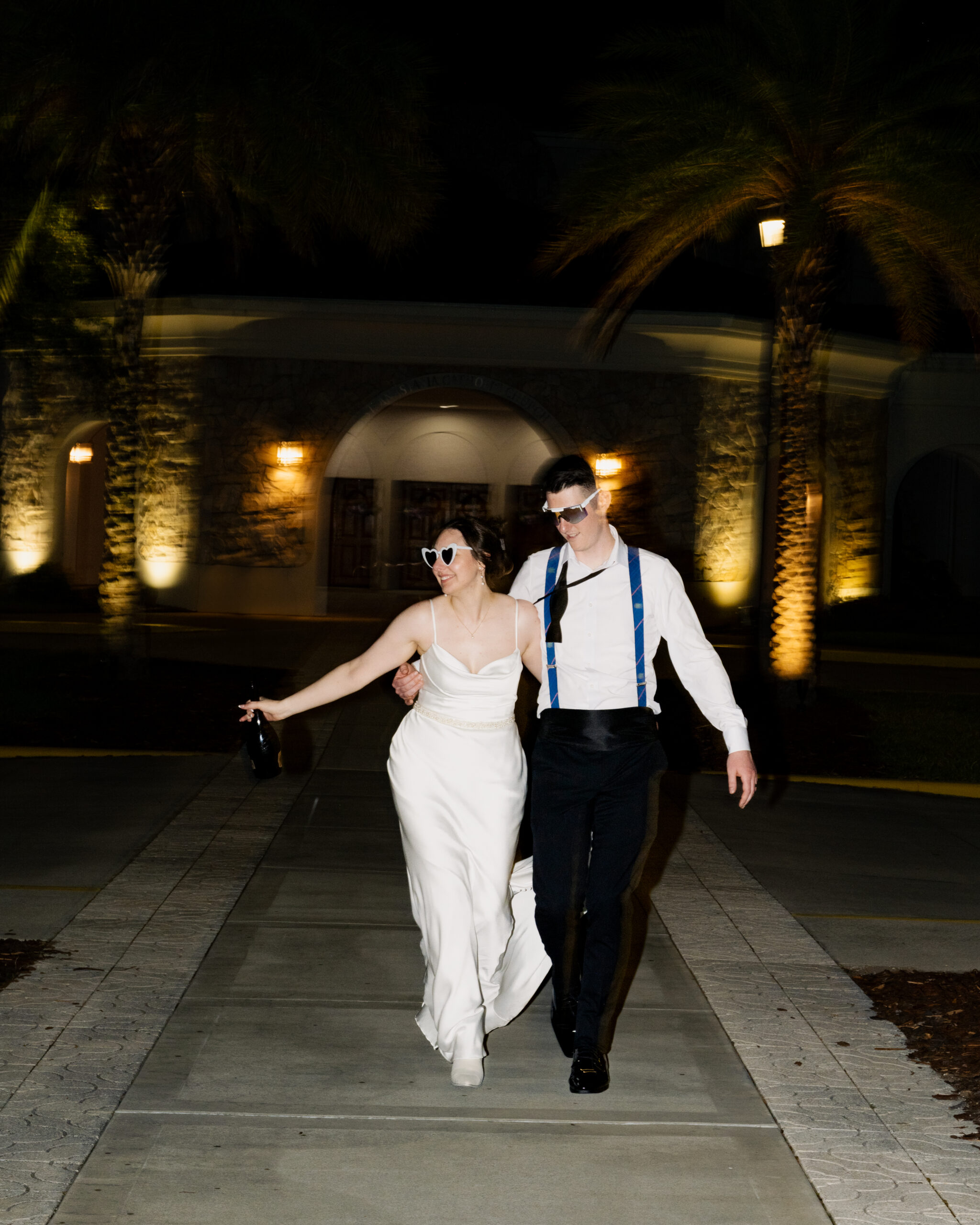 Newlyweds walk arm-in-arm laughing together in front of palm trees and the lit facade of St. Anastasia Catholic Church in St. Augustine, Florida during nighttime wedding photos.
