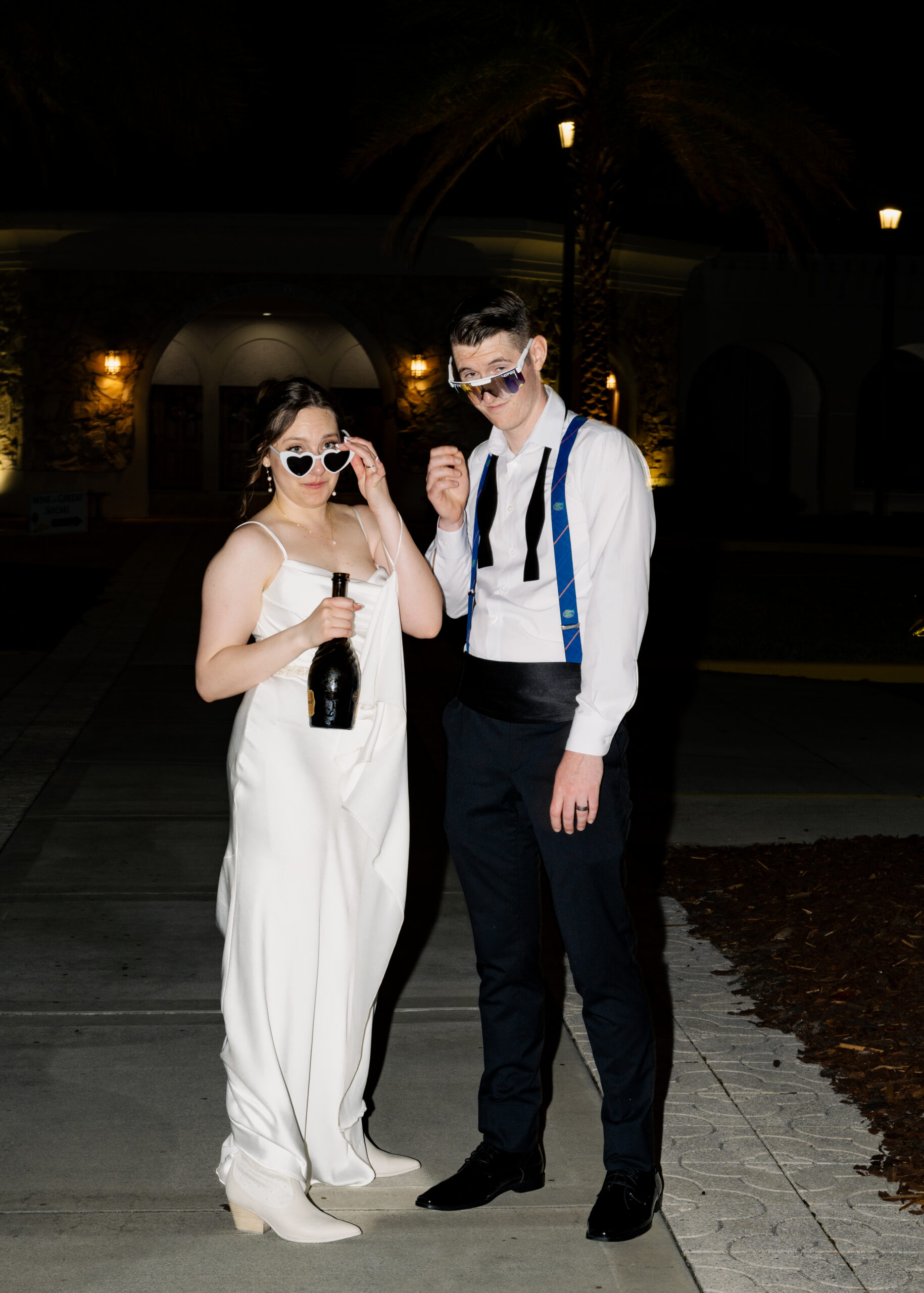 Bride and groom pose together under palm trees at night, holding a champagne bottle and wearing playful sunglasses at their St. Augustine Catholic wedding reception.