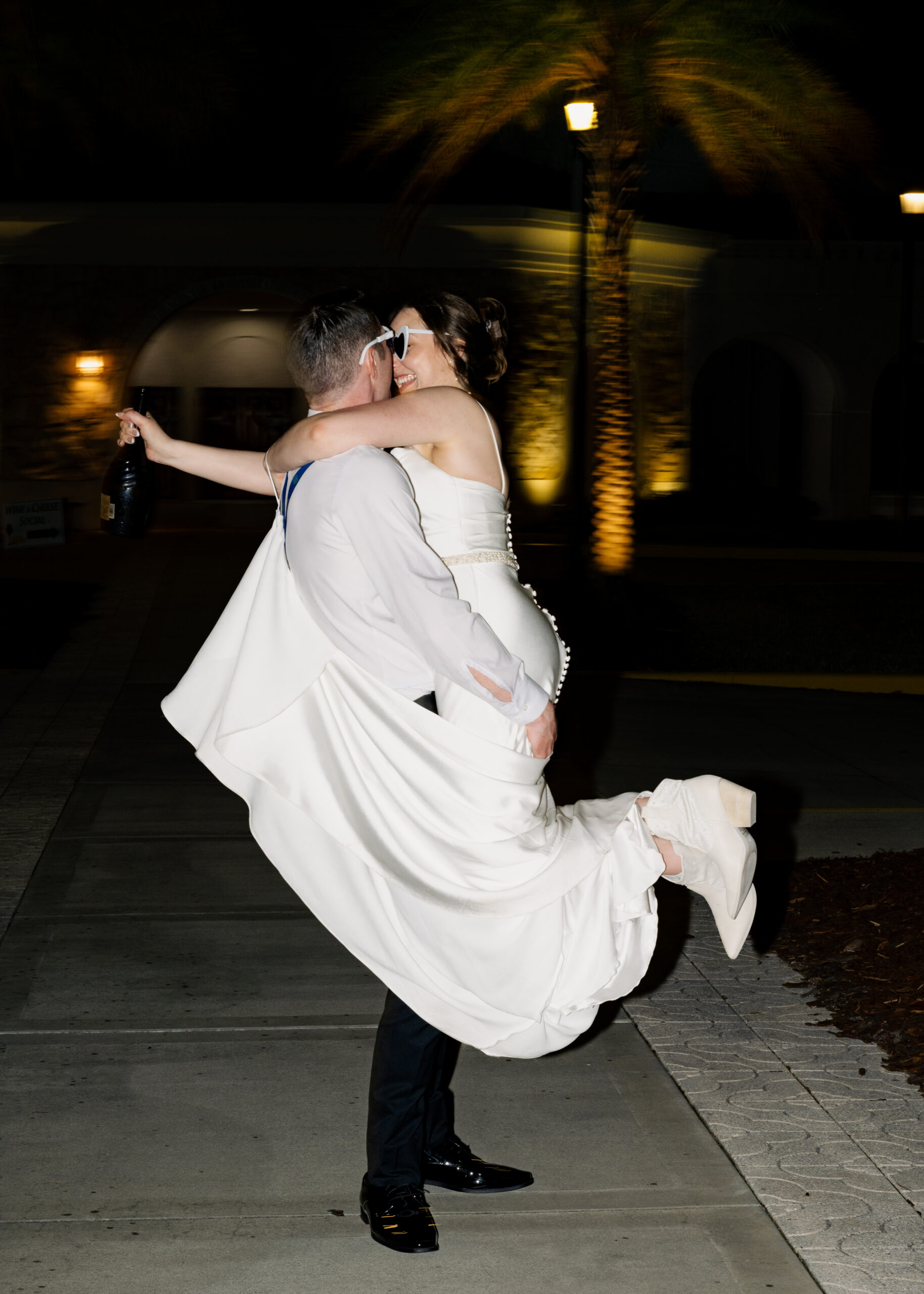Groom lifts the bride for a kiss during fun, late-night wedding portraits at St. Anastasia Catholic Church in St. Augustine, Florida, bride holding champagne bottle in hand.
